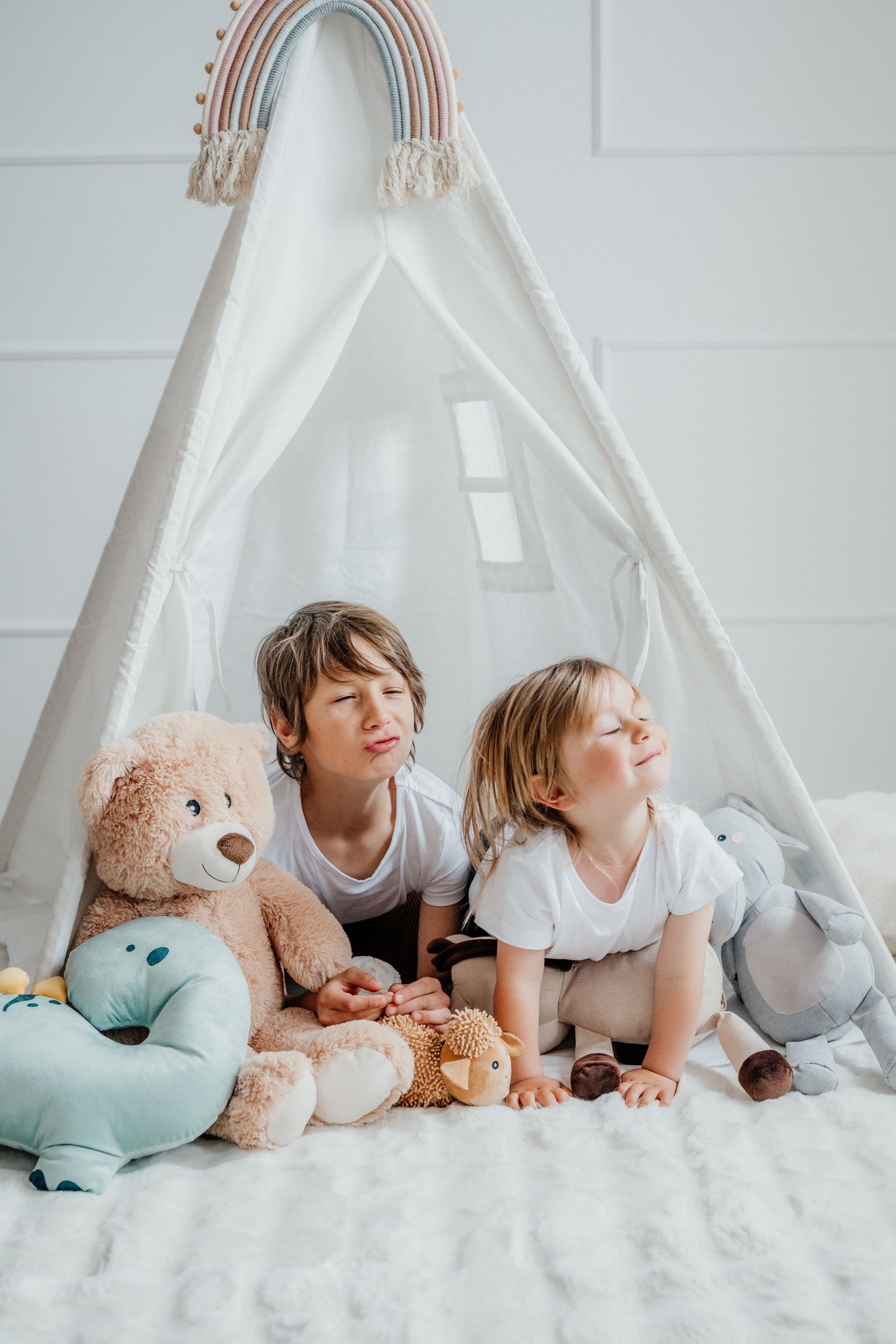 Two young boys sit together inside a white teepee tent surrounded by stuffed animals on a fluffy white rug.