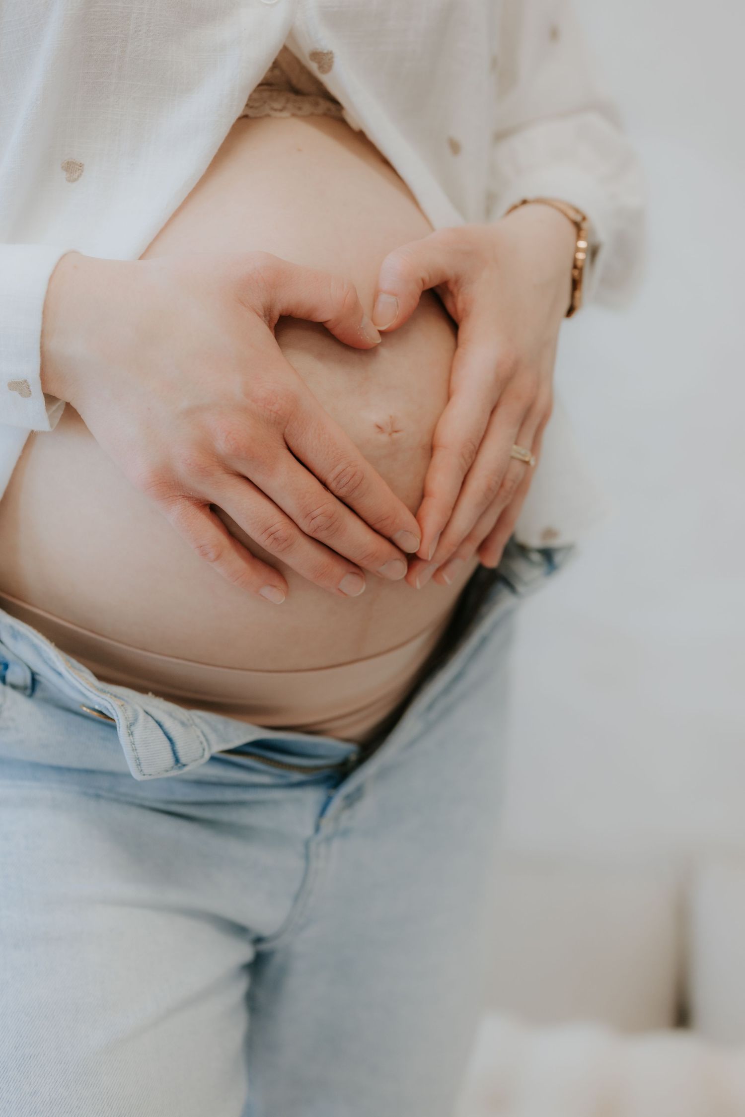 Close-up of pregnant woman making heart shape with hands on her bare baby bump in jeans.