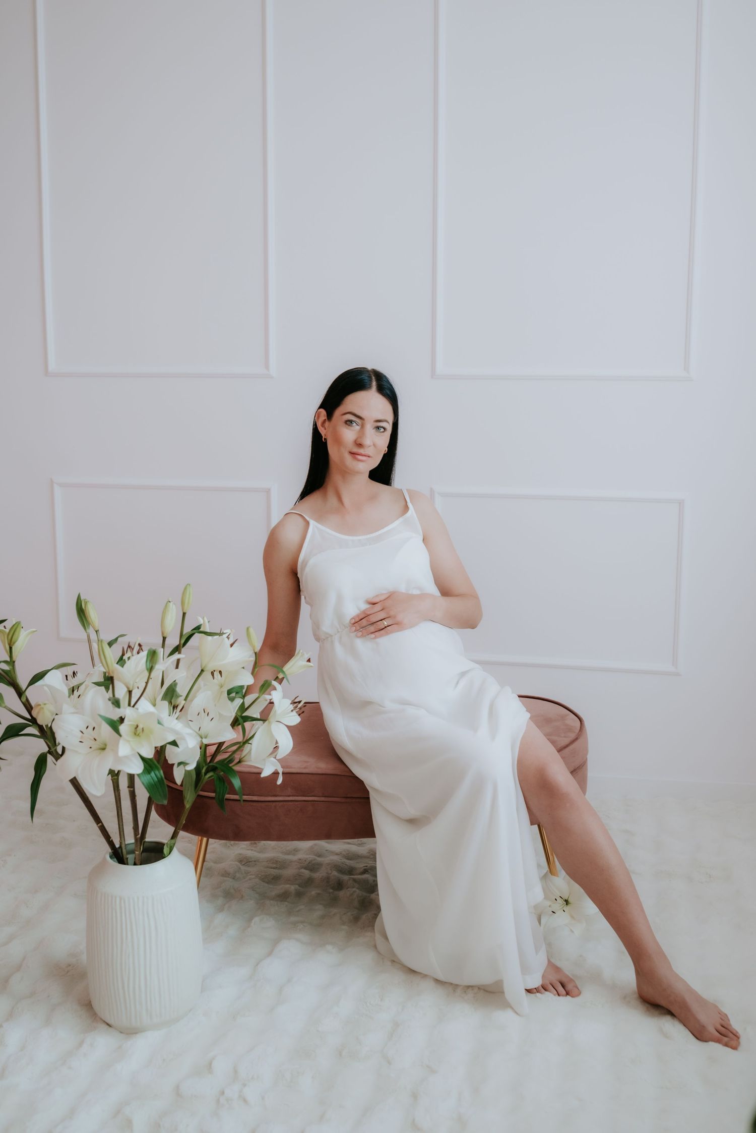 Pregnant woman in white slip dress seated on bench beside white lily vase arrangement.