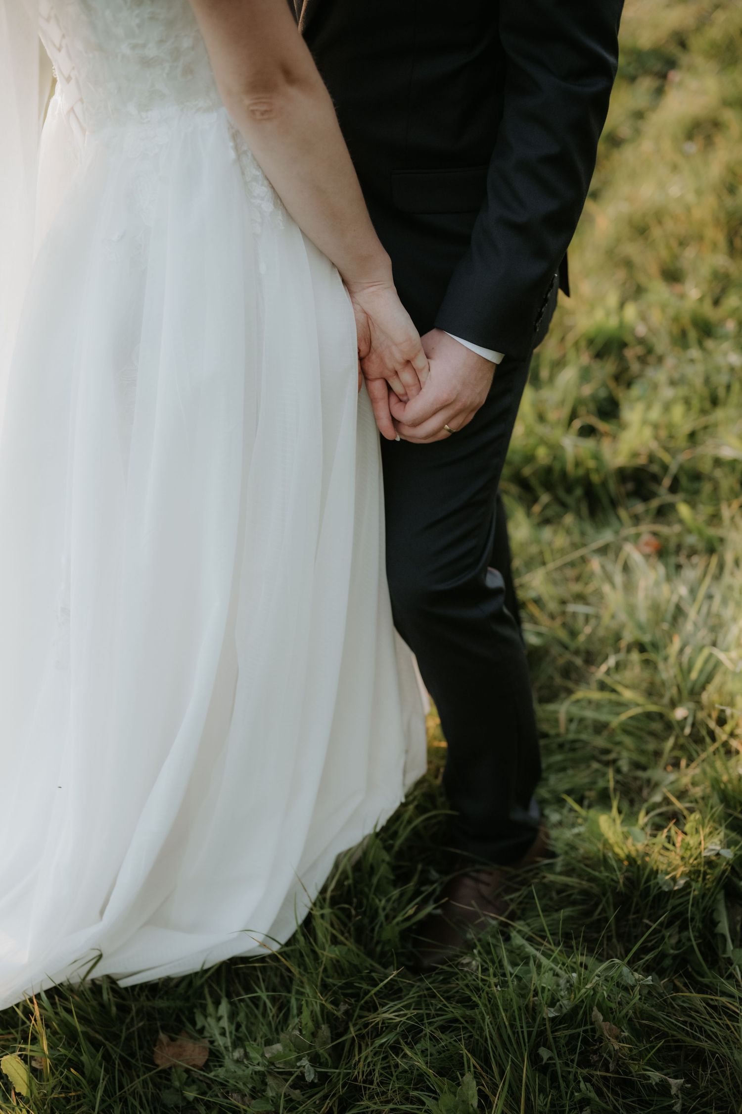Bride and groom holding hands outdoors, wearing white wedding dress and black suit, standing on green grass.