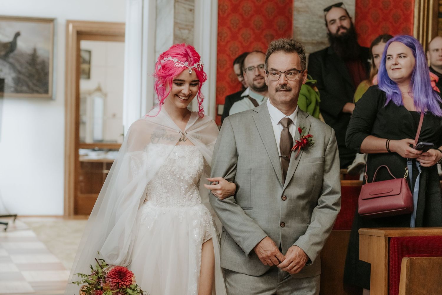 Bride with pink hair in white gown walks with suited groom at wedding ceremony, guests watching in elegant red-walled venue.