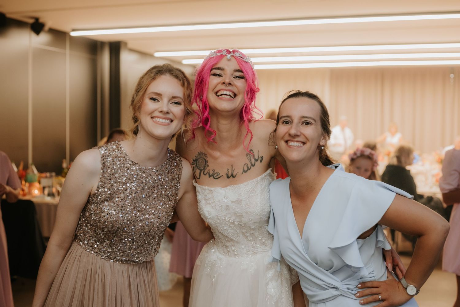 Bride with pink hair in white dress poses with two smiling women in formal attire at a wedding reception.