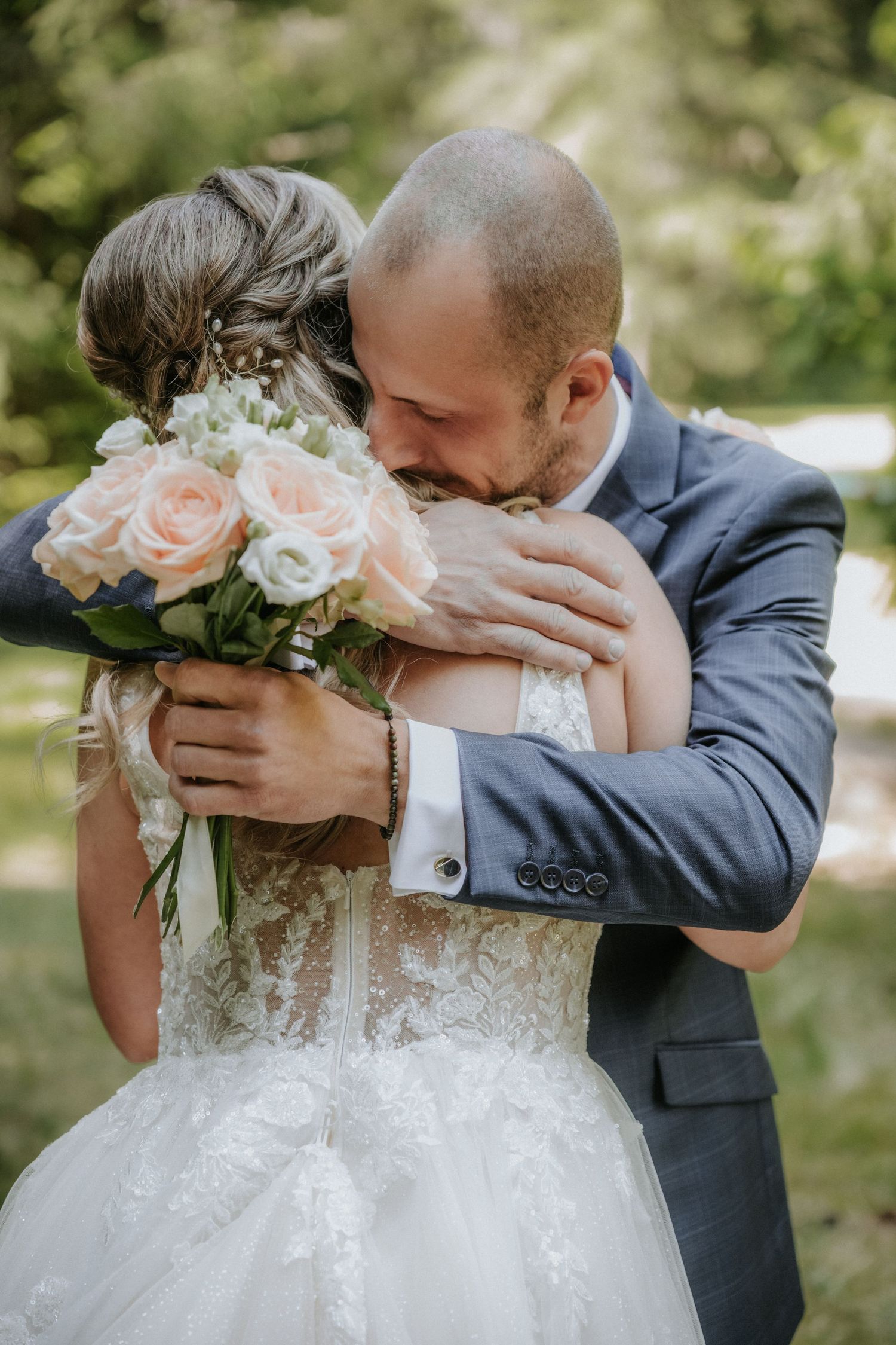 Groom in blue suit embracing bride in lace wedding dress, holding pink and white rose bouquet outdoors.