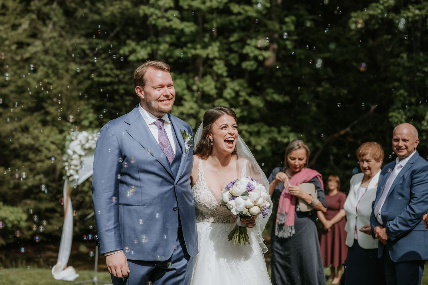 Joyful newlyweds walk down the aisle outdoors, bride holding bouquet, guests celebrating with bubbles.