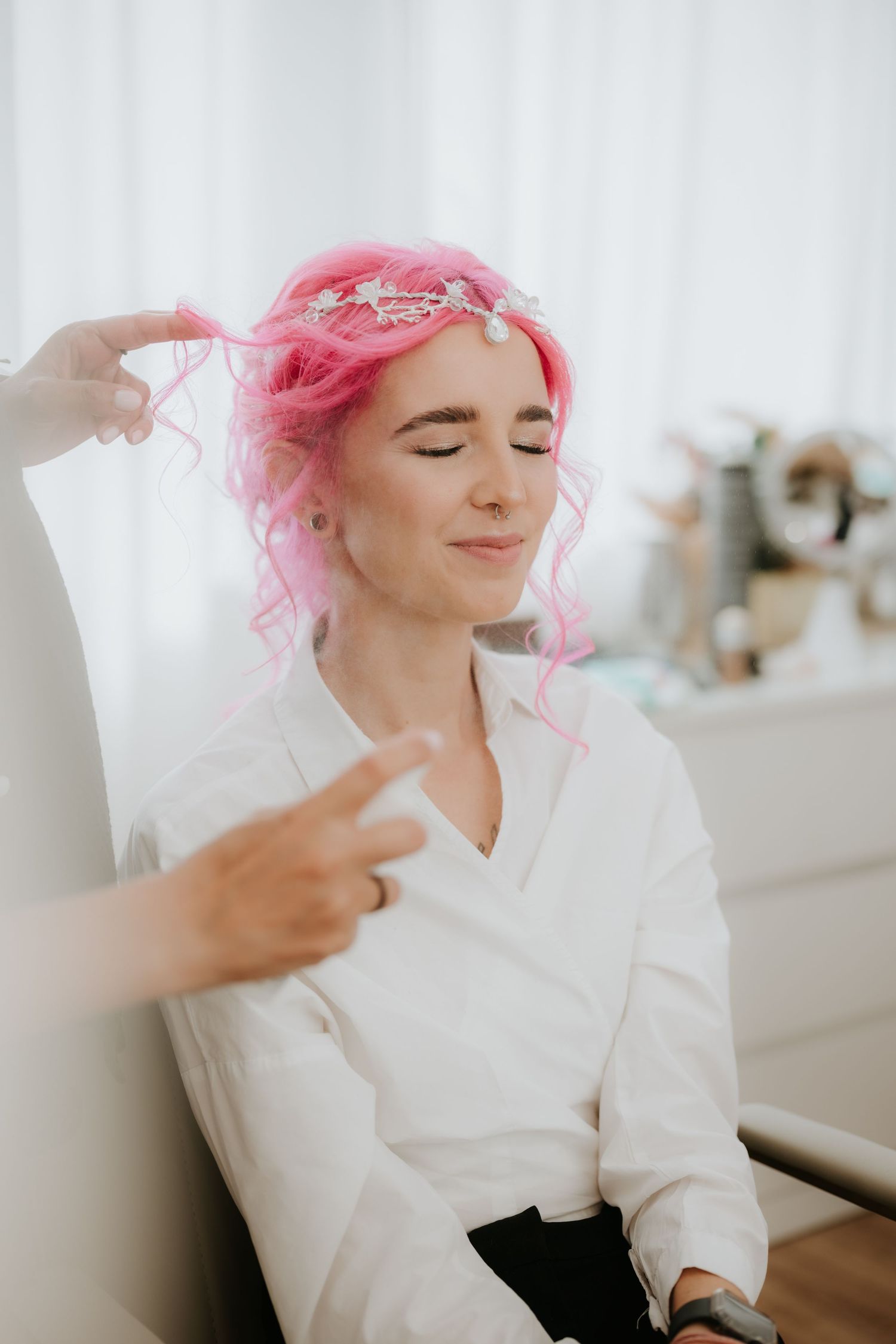 Woman with pink curly hair and floral crown gets styled by hairdresser in bright salon setting.