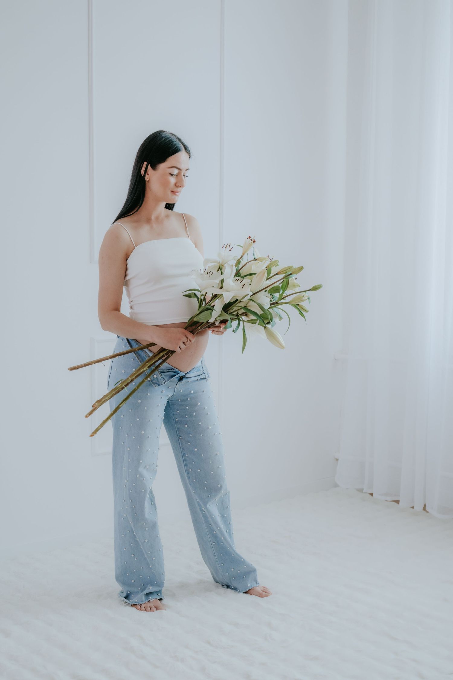 Pregnant woman in white crop top and pearl jeans holding a bouquet of white lilies.