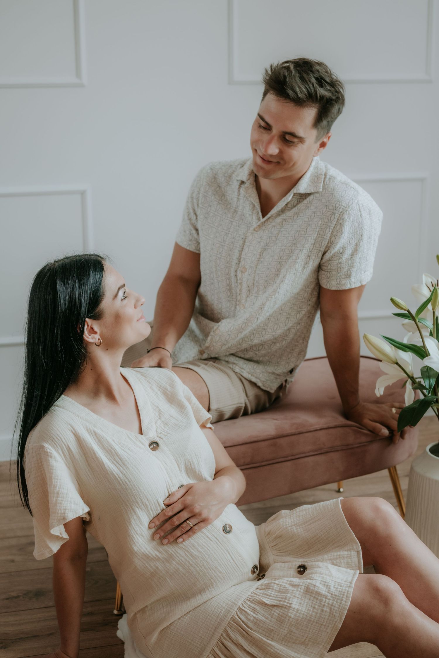 Couple sharing tender moment, pregnant woman in cream dress seated on floor, man on pink bench.
