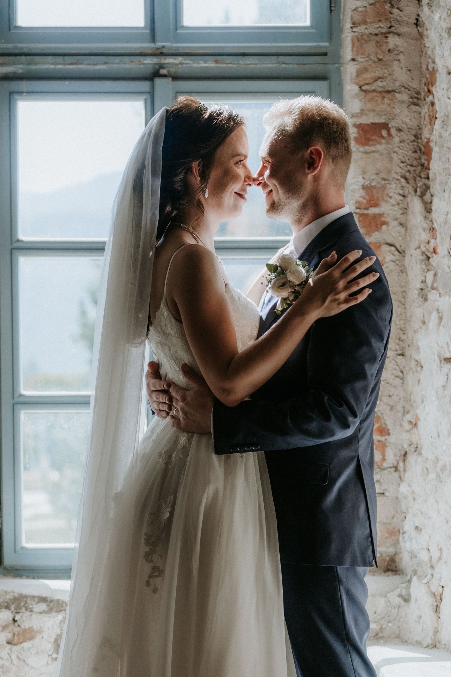Bride and groom in elegant wedding attire share a romantic moment, touching foreheads near a rustic brick wall window.