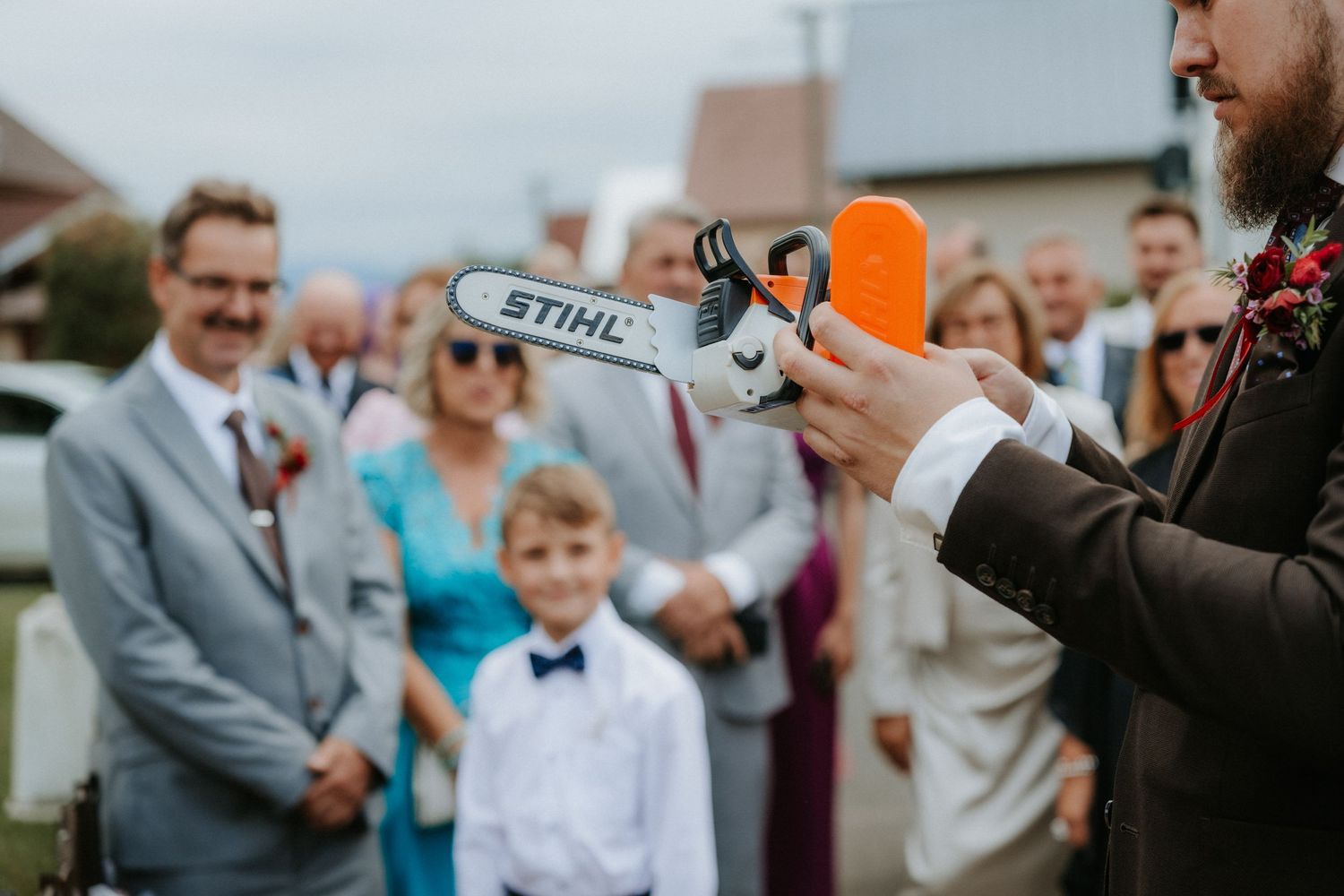 Groom holds a miniature STIHL chainsaw toy at a wedding, with guests smiling in the background.