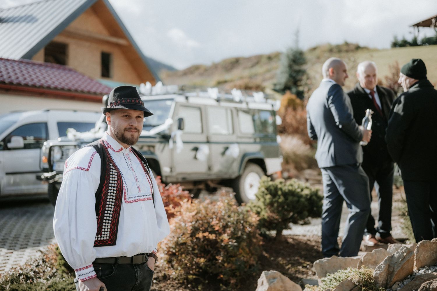 Man in traditional Eastern European folk costume stands outdoors near a white SUV with guests conversing in background.