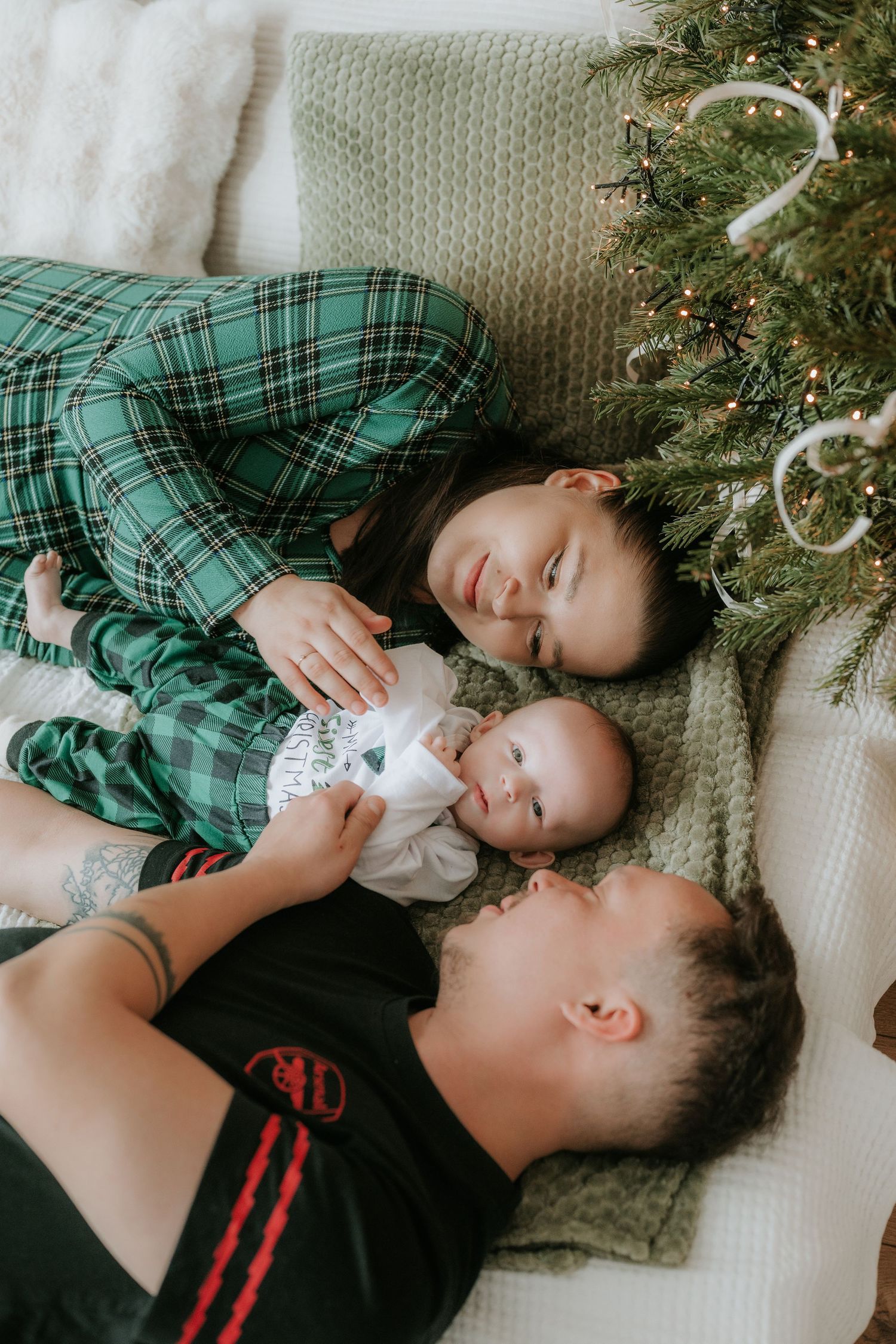 Family lying near Christmas tree with parents in plaid pajamas cuddling newborn baby on white blanket.