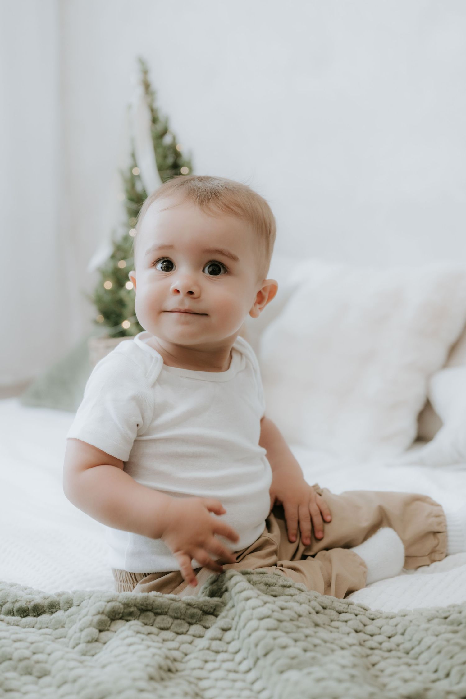 Adorable baby in white onesie sitting on green knit blanket with Christmas tree in background.