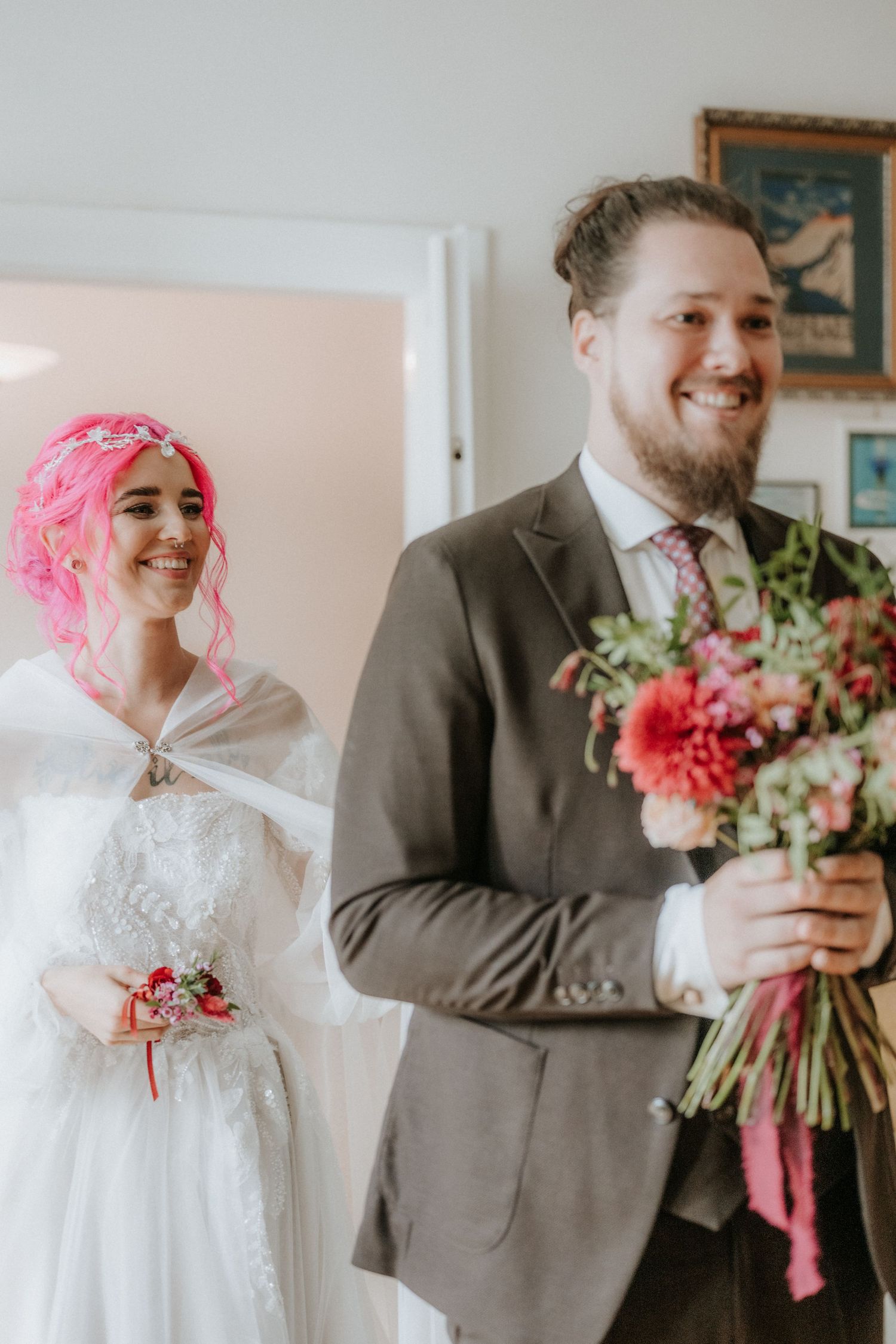 Groom in gray suit holds pink floral bouquet while bride with pink hair in white dress smiles behind him.