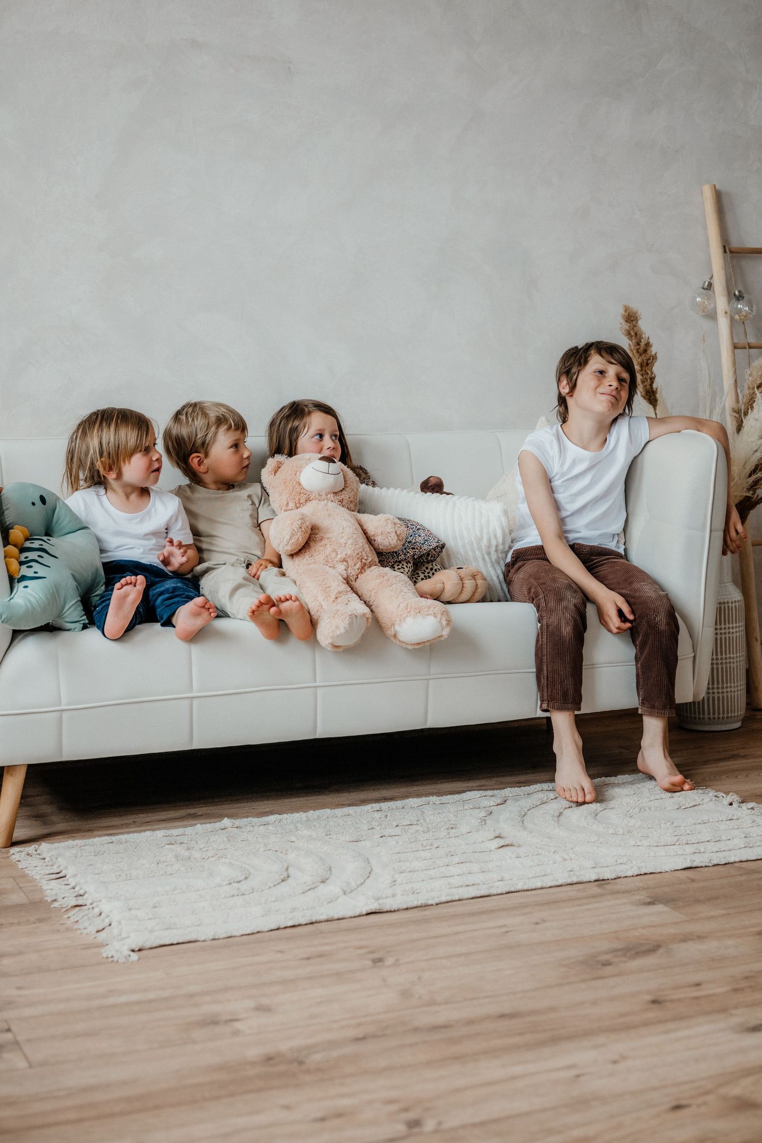 Four children sit together on a white sofa with a large teddy bear in a bright, cozy living room.
