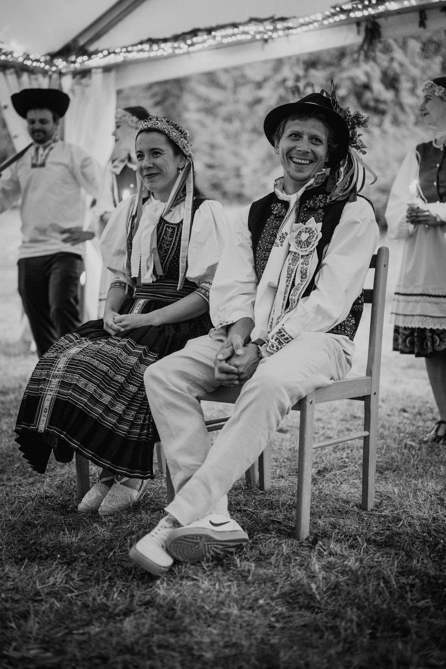 Black and white photo of a smiling couple in traditional folk costumes seated at an outdoor cultural event.