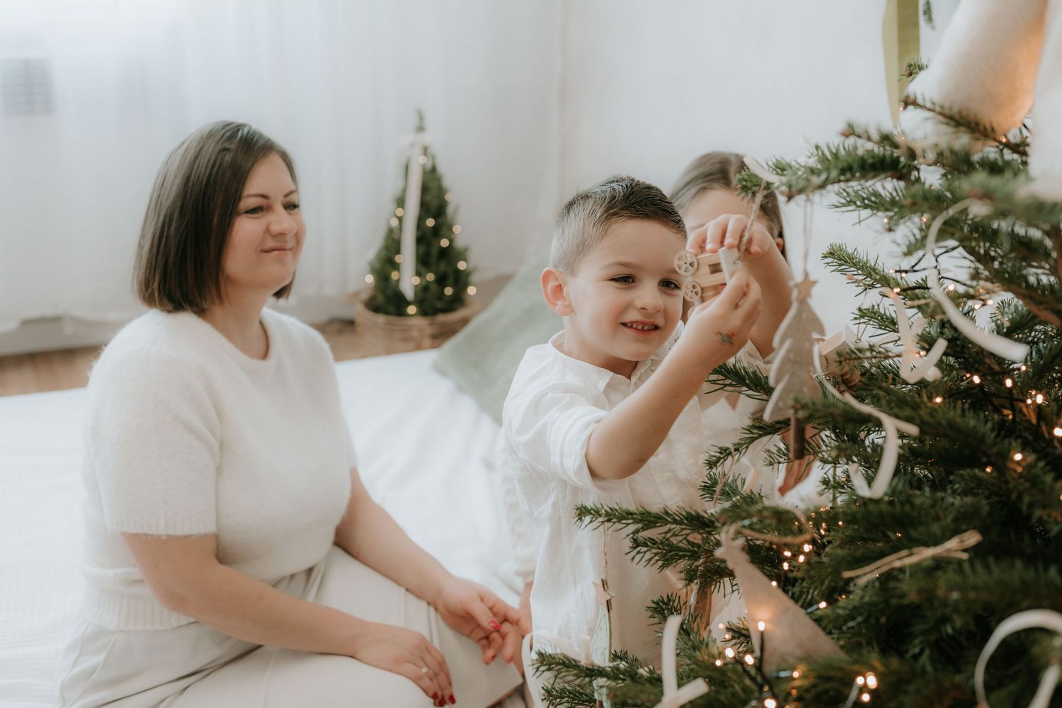 A young boy decorates a Christmas tree with ornaments while a woman in white watches warmly beside him.