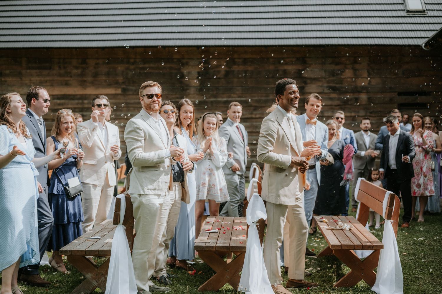 Two grooms in cream suits smile joyfully as wedding guests celebrate outdoors near a rustic barn.