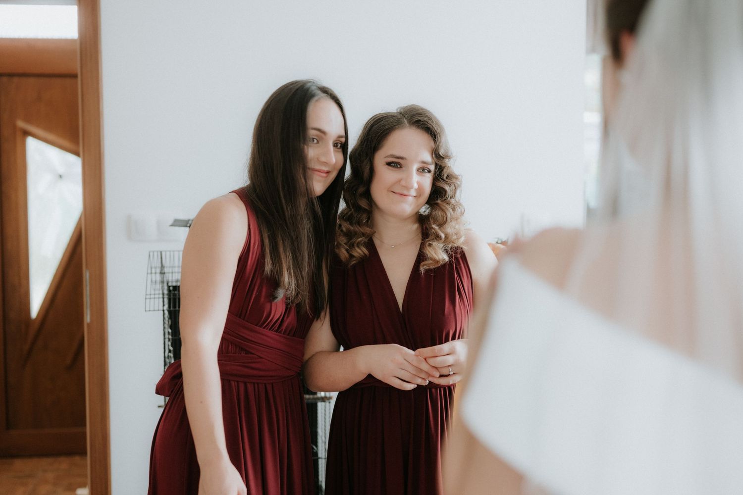 Two bridesmaids in burgundy dresses admire a wedding gown during bridal preparations.