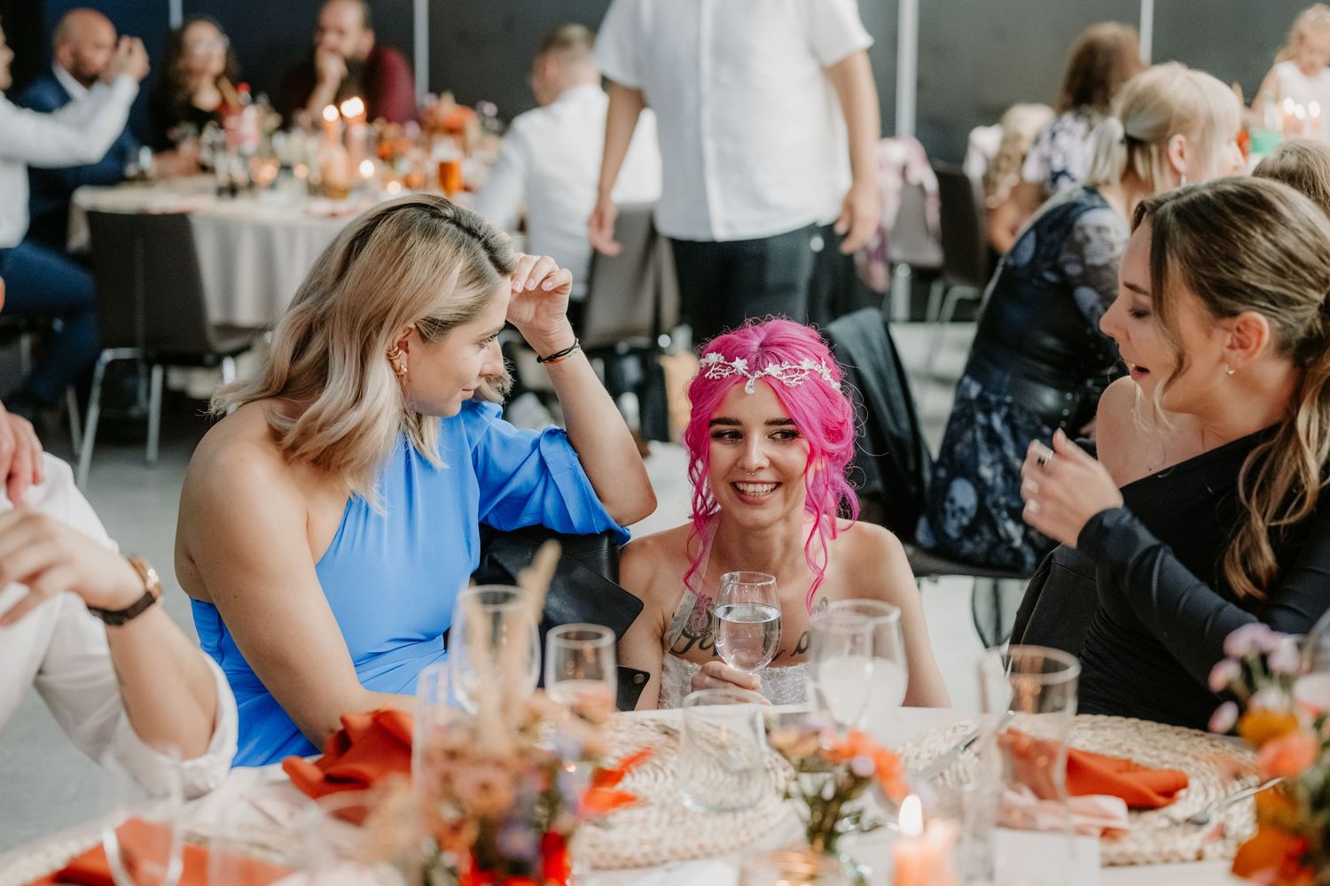 Women laughing together at a wedding reception table, one with pink hair holding a wine glass.