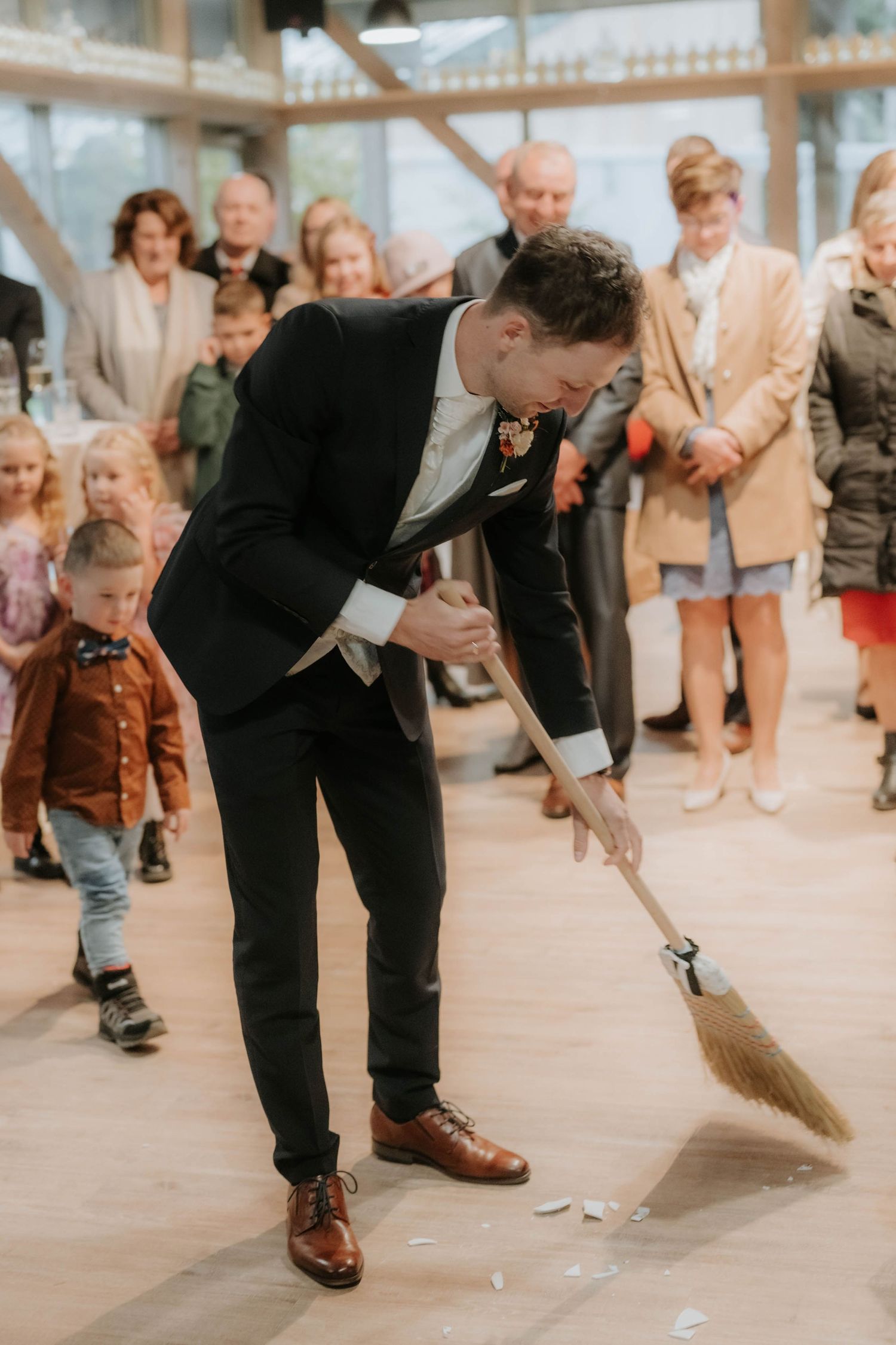Groom in black suit sweeping floor with broom at wedding ceremony while guests watch in elegant venue.