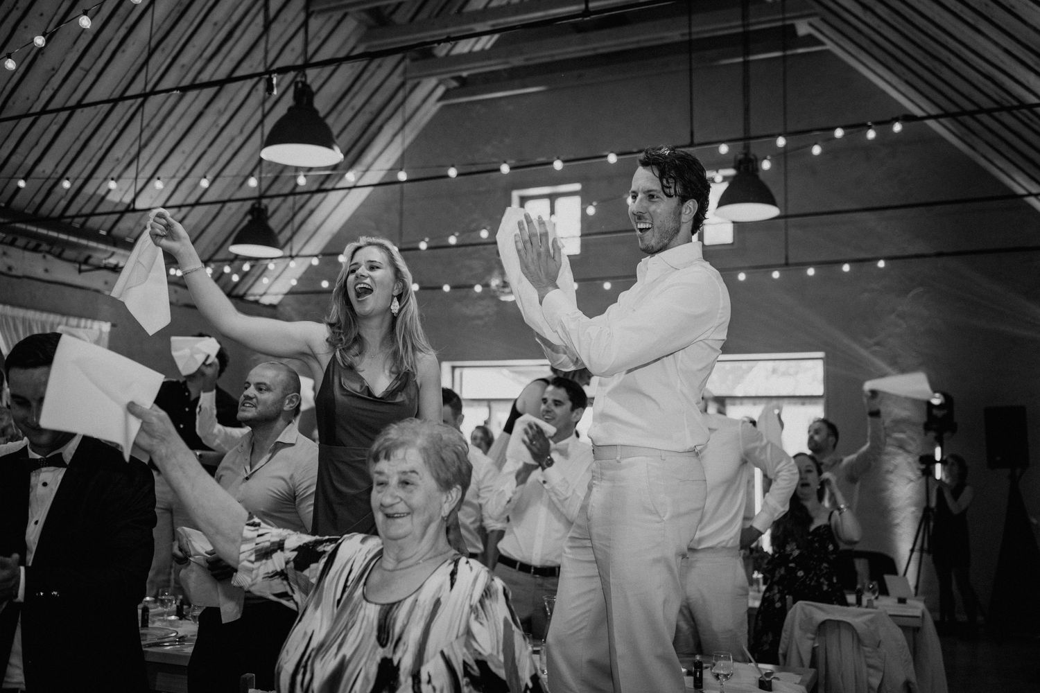 Black and white photo of joyful wedding guests dancing and celebrating at a festive indoor reception venue.