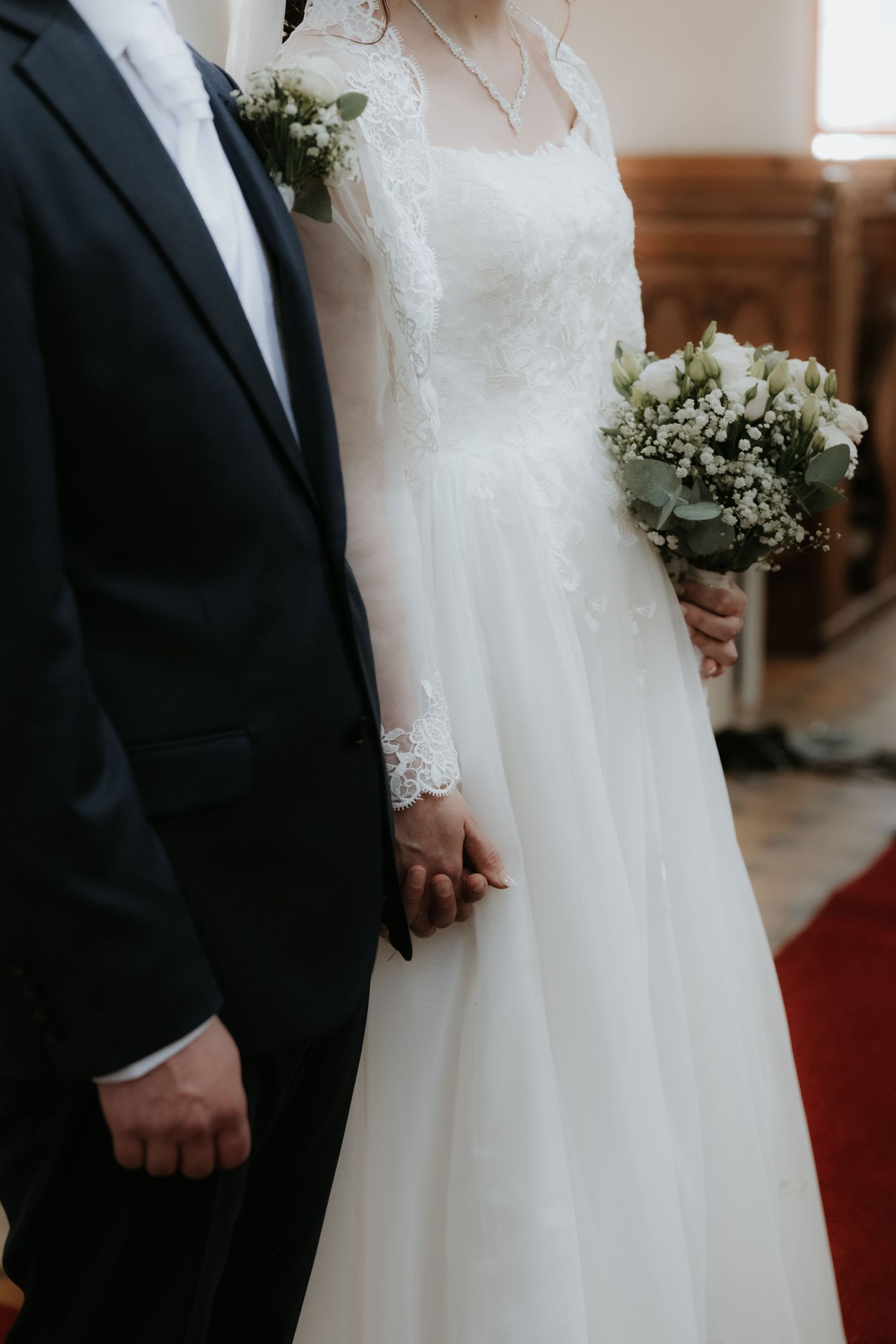 Bride in lace wedding gown holding white bouquet stands beside groom in dark suit during church ceremony.
