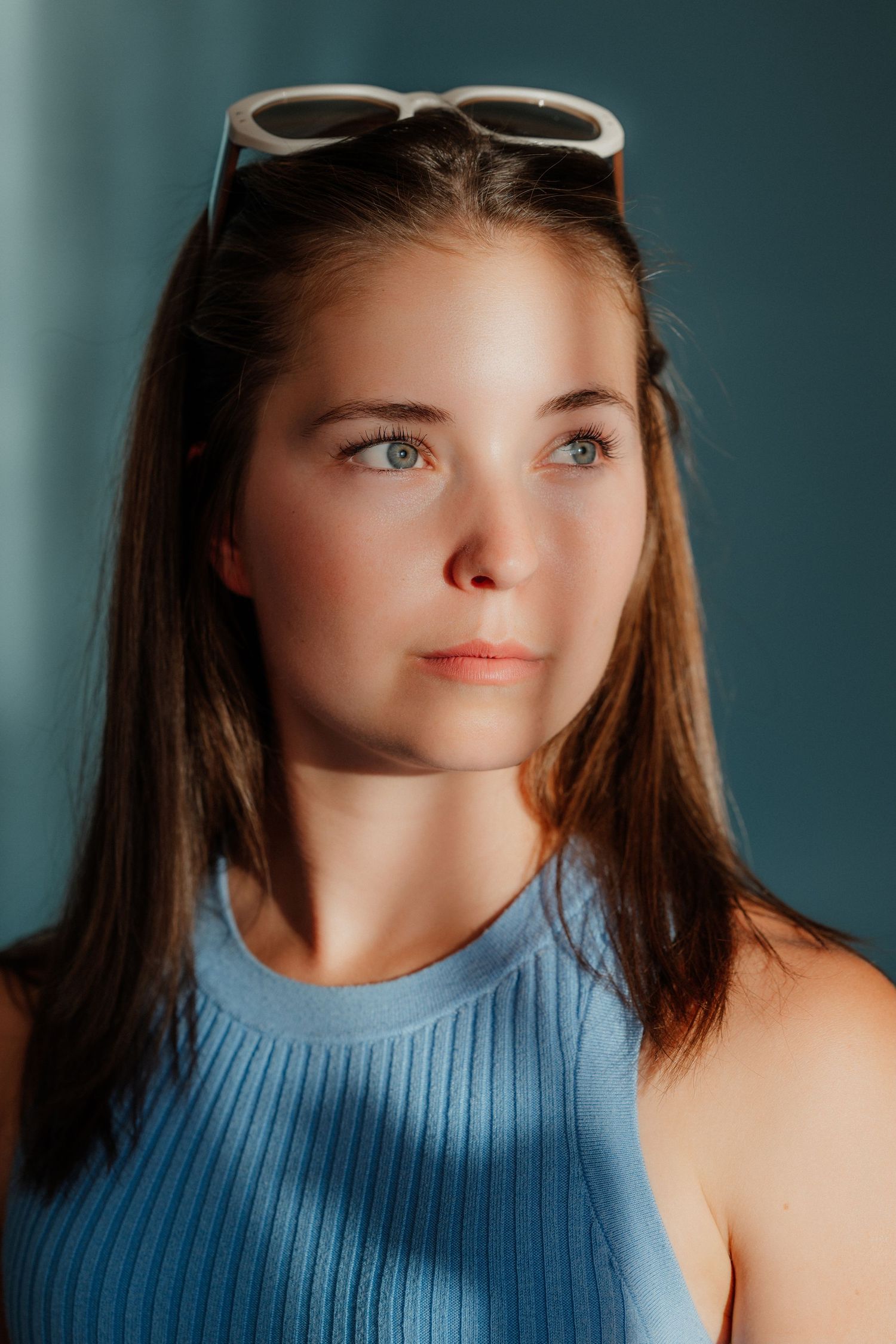 Young woman with blue eyes and white sunglasses on her head wearing a blue ribbed tank top in warm sunlight.