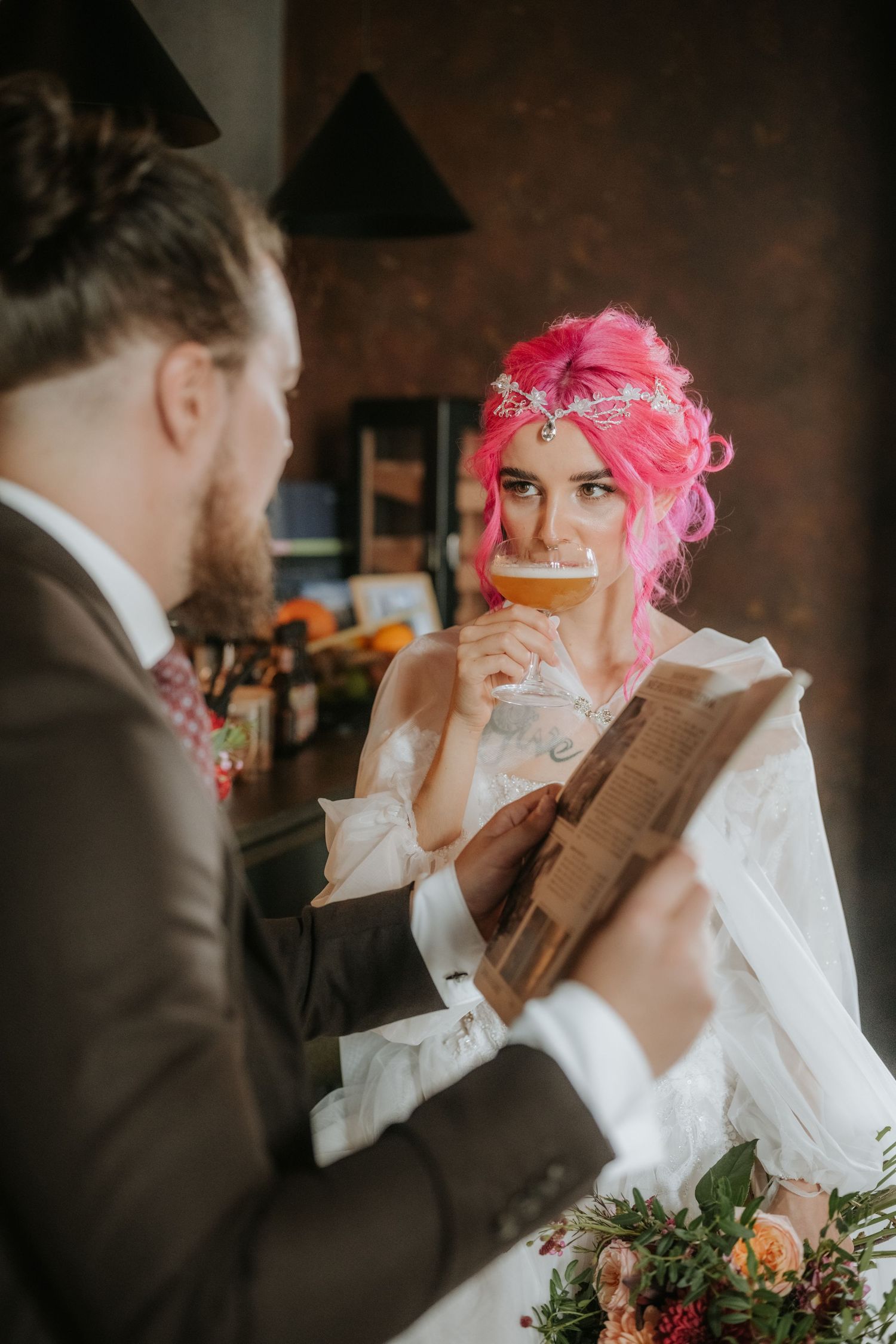 Bride with pink hair sipping beer while groom reads vows at unconventional wedding ceremony.