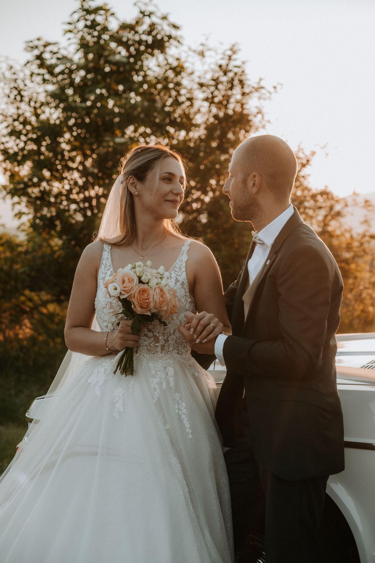 Bride and groom share a romantic sunset moment, she holds a peach rose bouquet in a lace gown beside a white car.