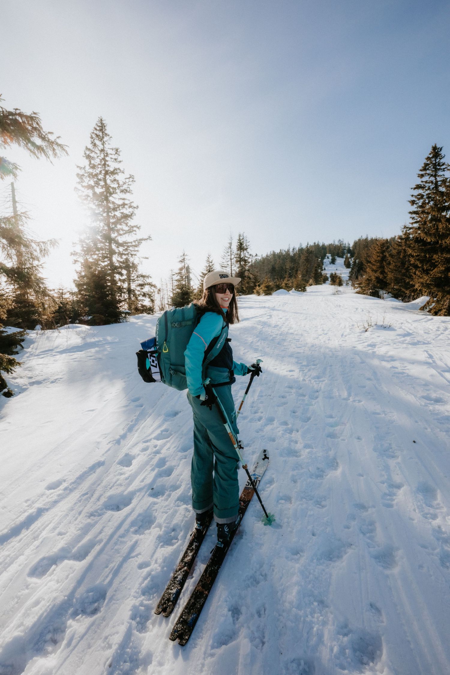 Female skier in teal jumpsuit posing with poles on sunny snowy mountain trail.