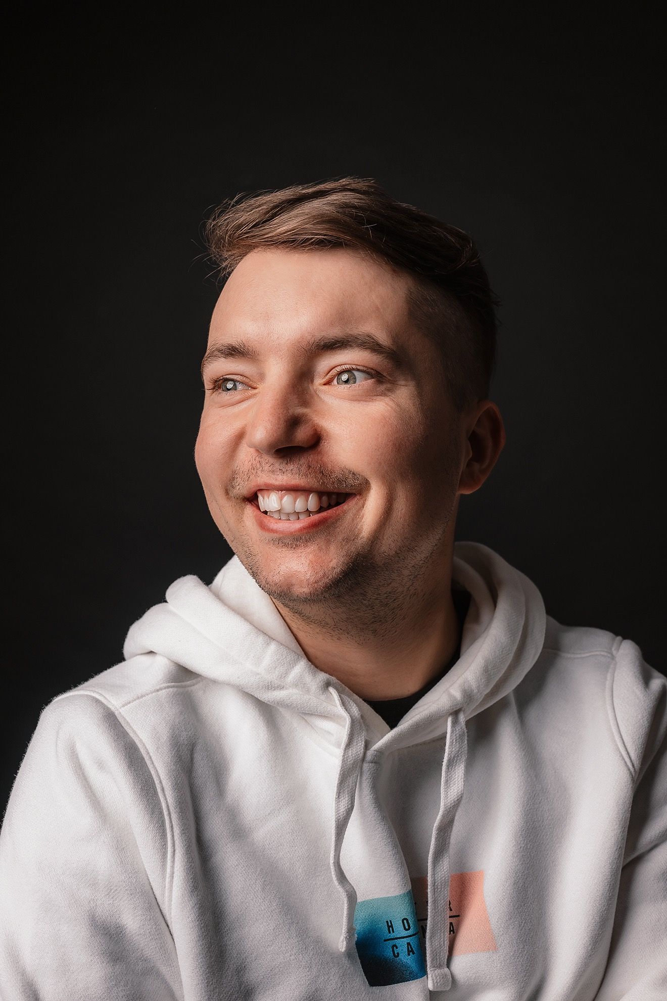 Smiling young man in white hoodie posing against dark background in studio portrait.