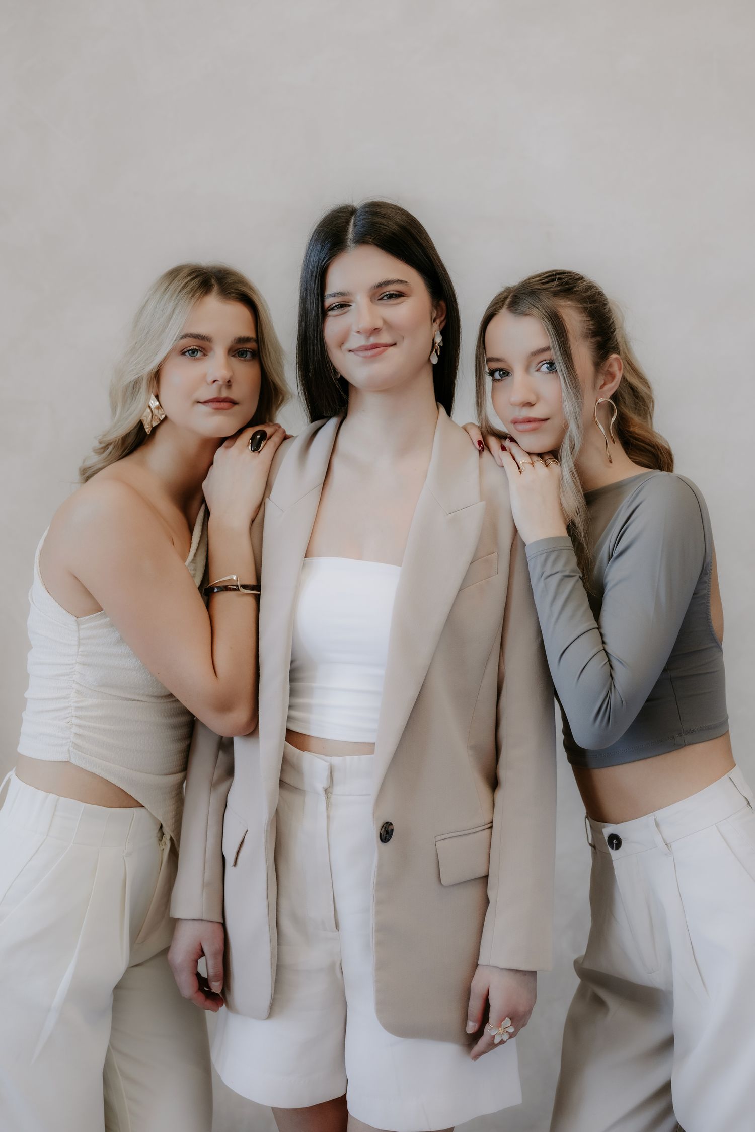 Three fashionable women in neutral tones posing together, center wearing beige blazer and white crop top.