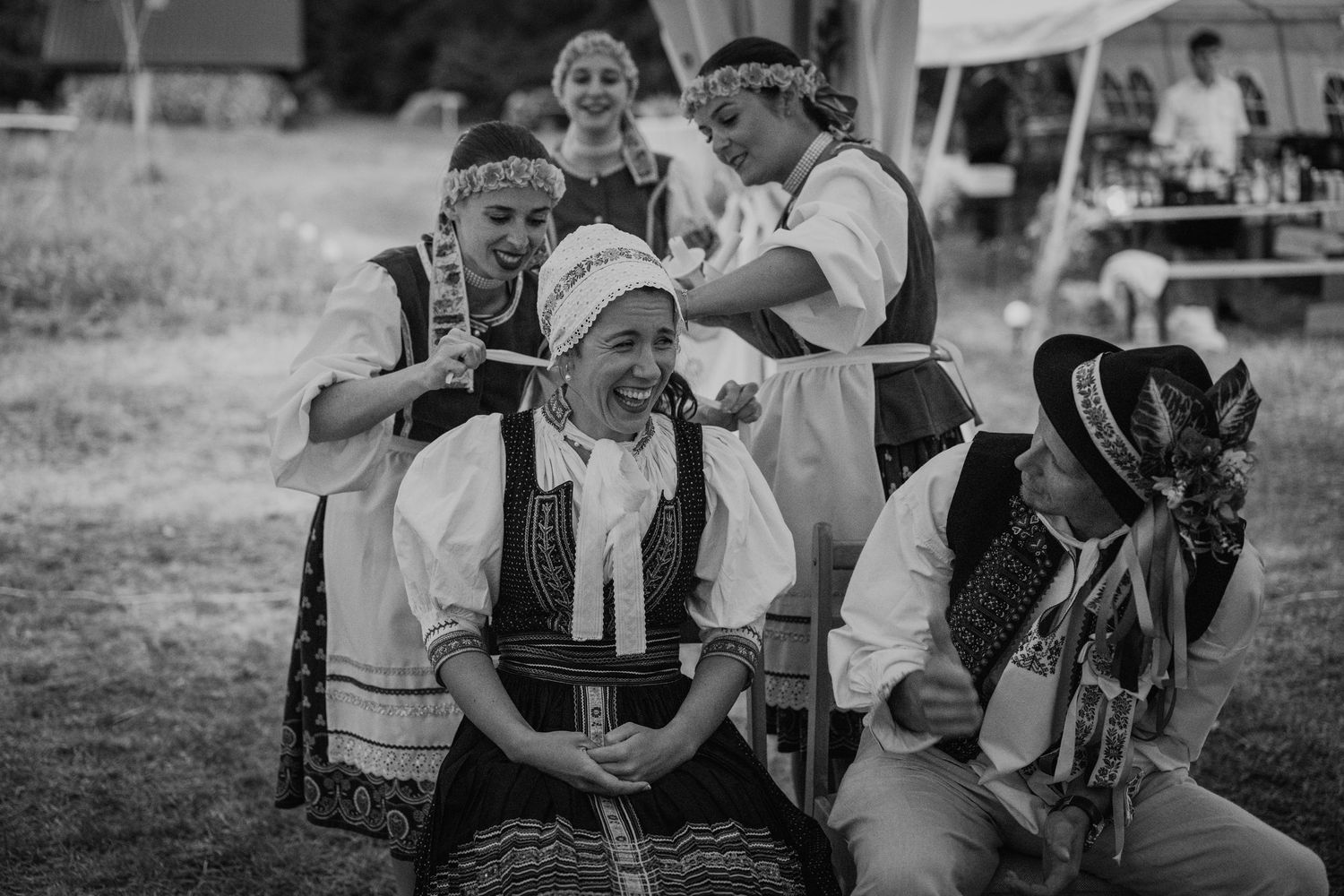 Black and white photo of people in traditional folk costumes laughing together at an outdoor cultural festival.
