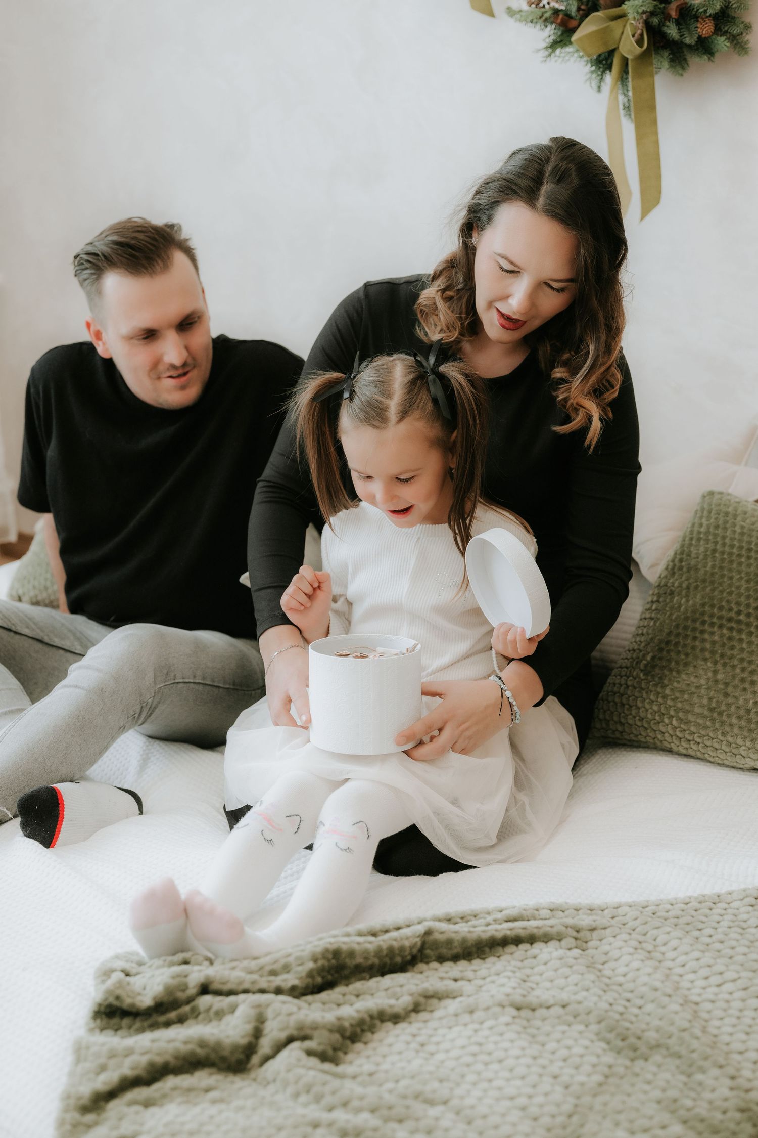 Loving family with toddler girl exploring Christmas gift box on cozy white bed.
