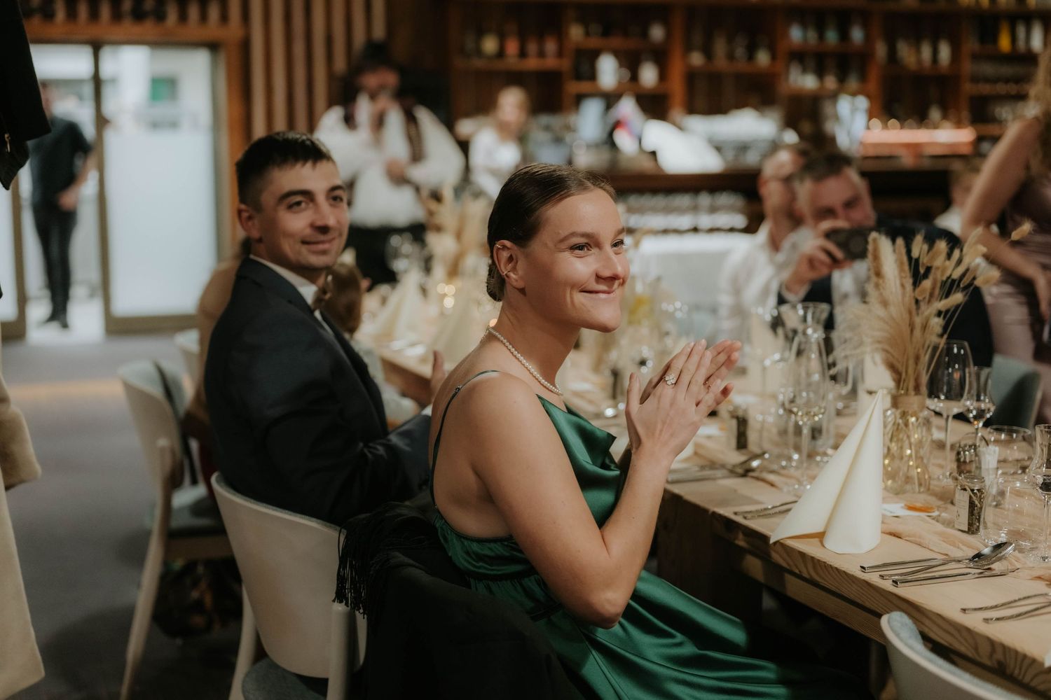 Guests in formal attire applaud at an elegant restaurant dinner event, woman in green dress smiling.