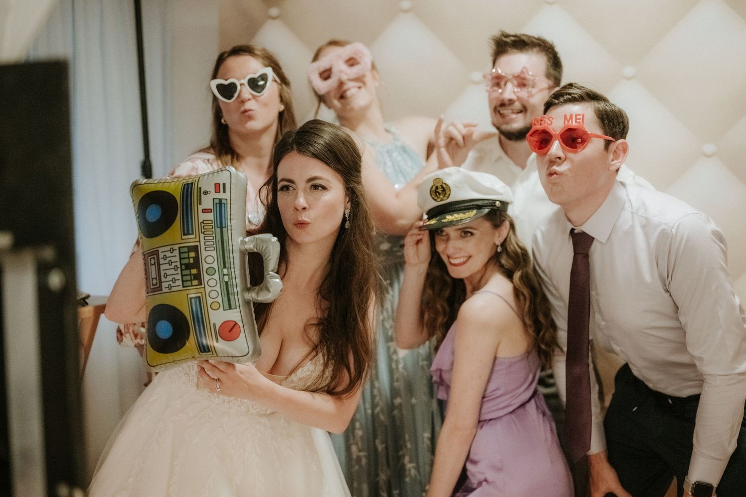 Wedding party posing with inflatable boombox and fun props including heart sunglasses and captain hat.