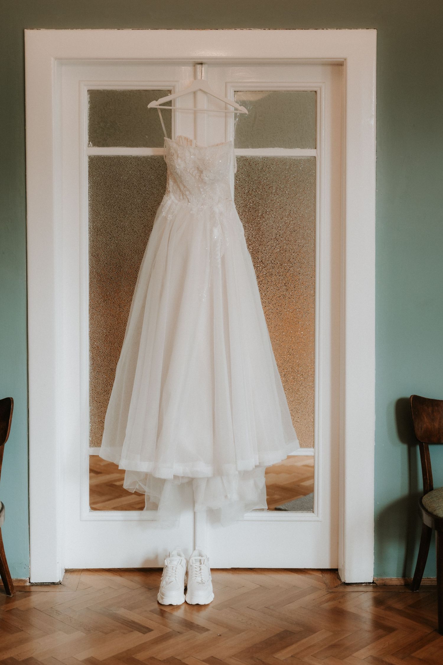 White lace A-line wedding dress hanging on door frame with matching bridal shoes below, elegant bridal preparation photo.