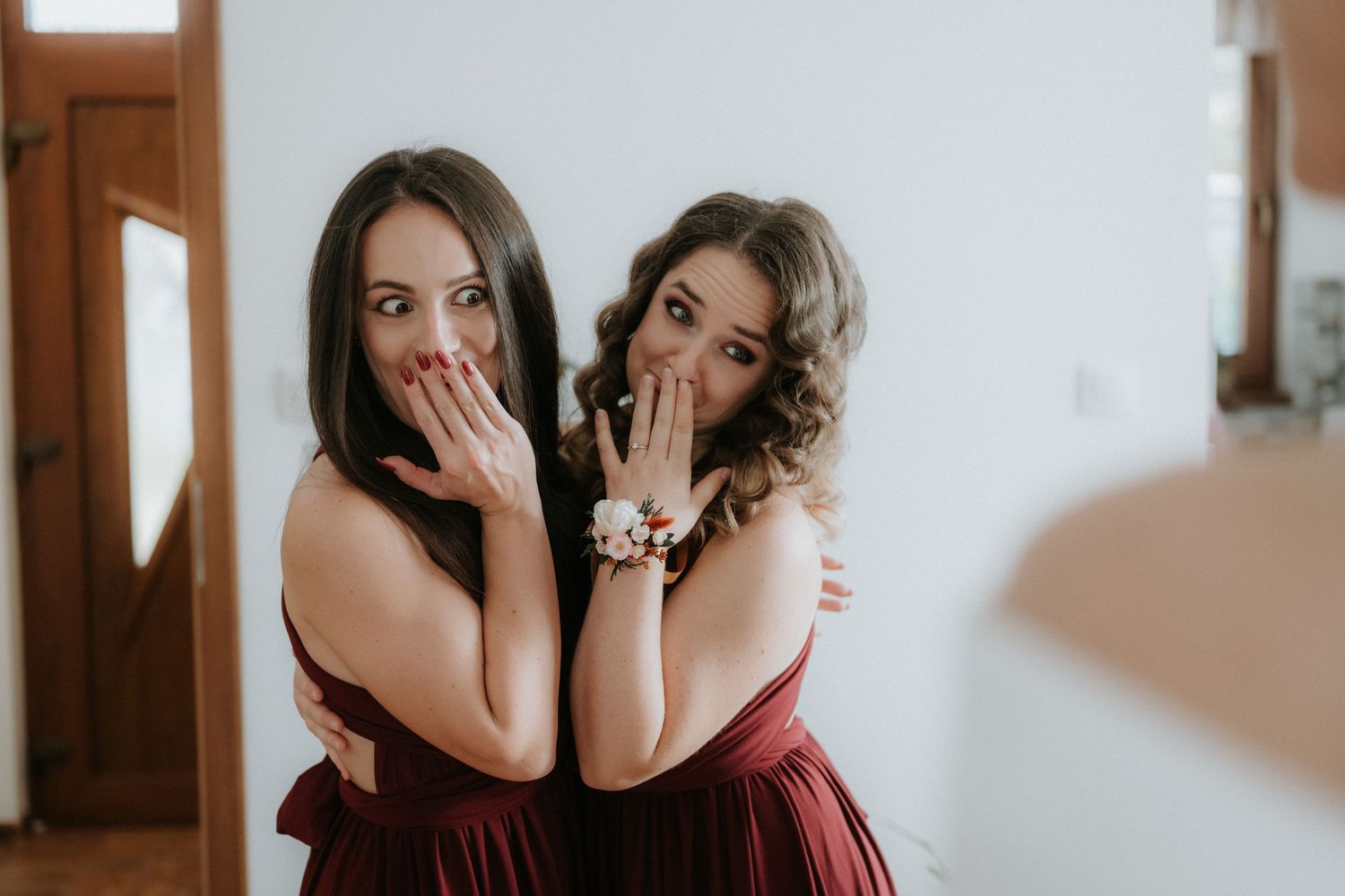 Two bridesmaids in burgundy dresses posing playfully with hands over mouths showing wrist corsages.