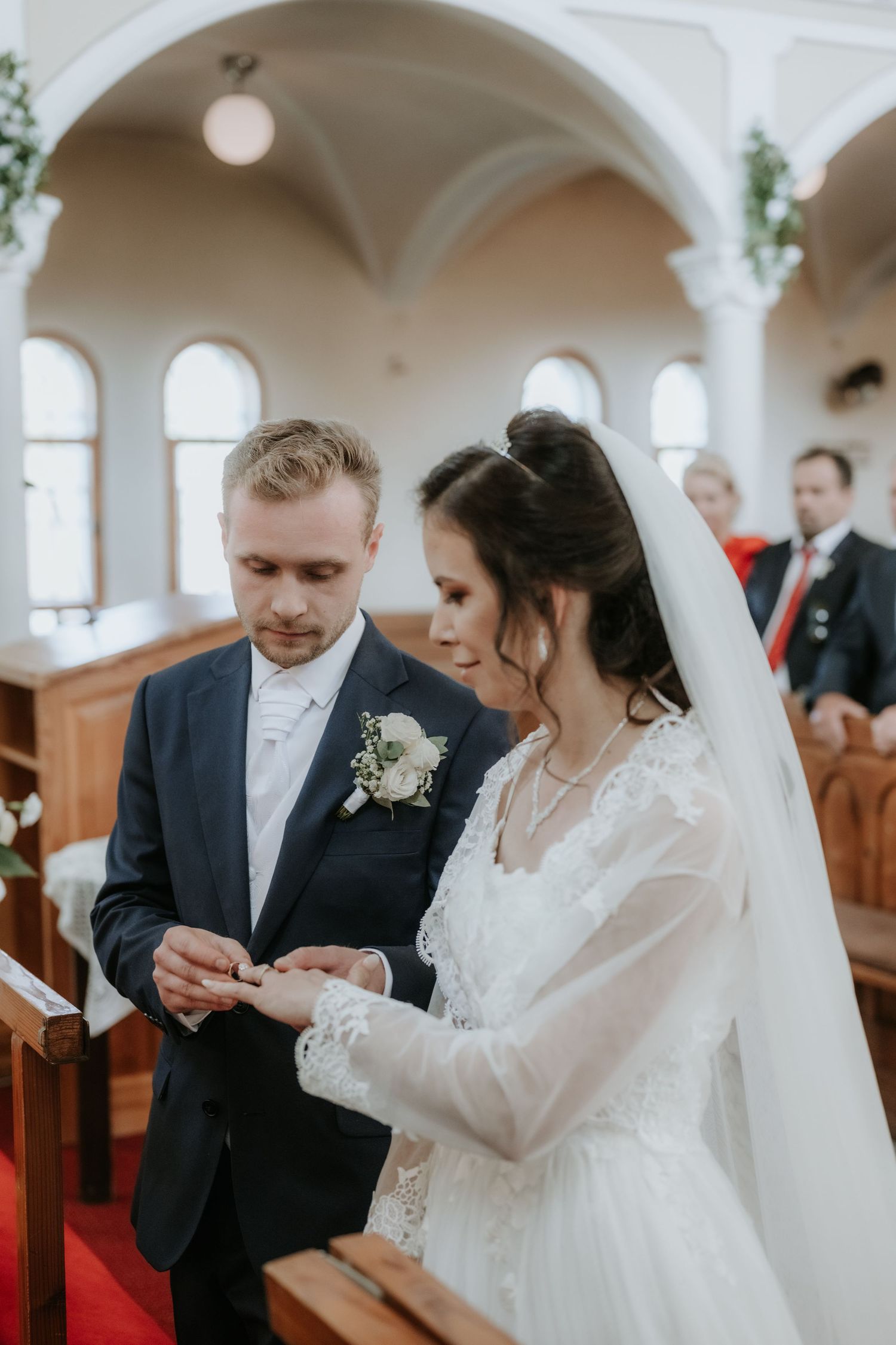 Bride and groom exchange rings at church altar during wedding ceremony, groom in navy suit, bride in white lace gown with veil.