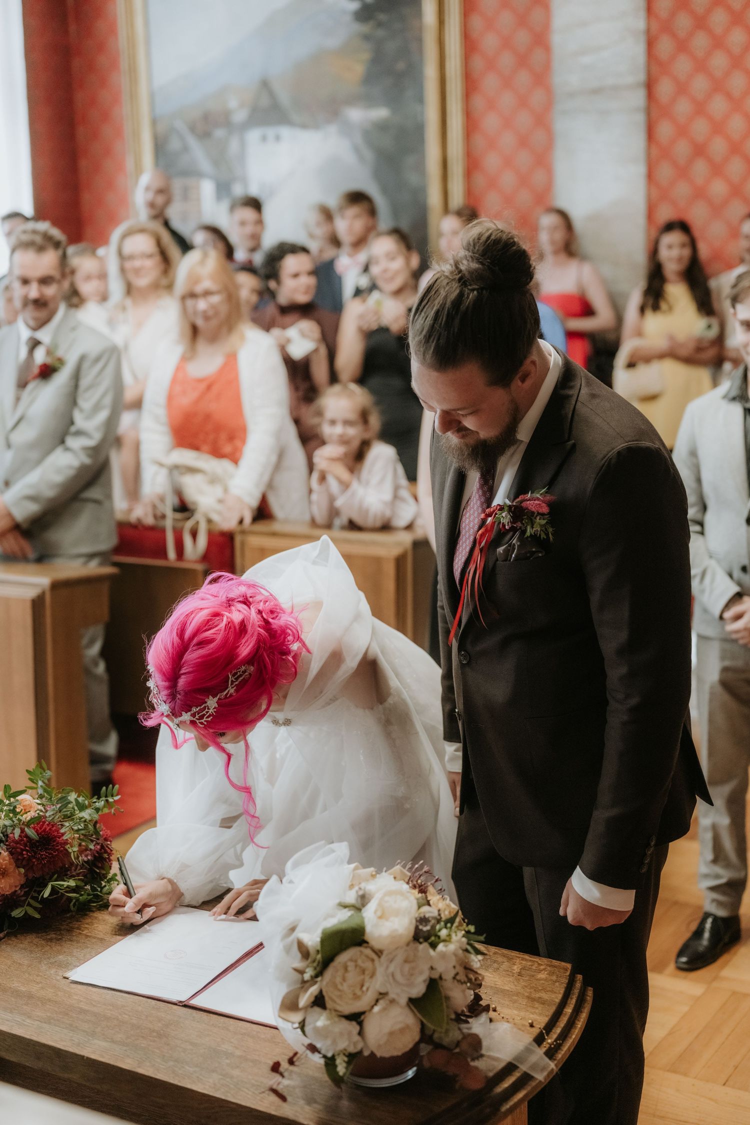 Bride with pink hair signing wedding register beside groom in black suit, guests watching in elegant hall.