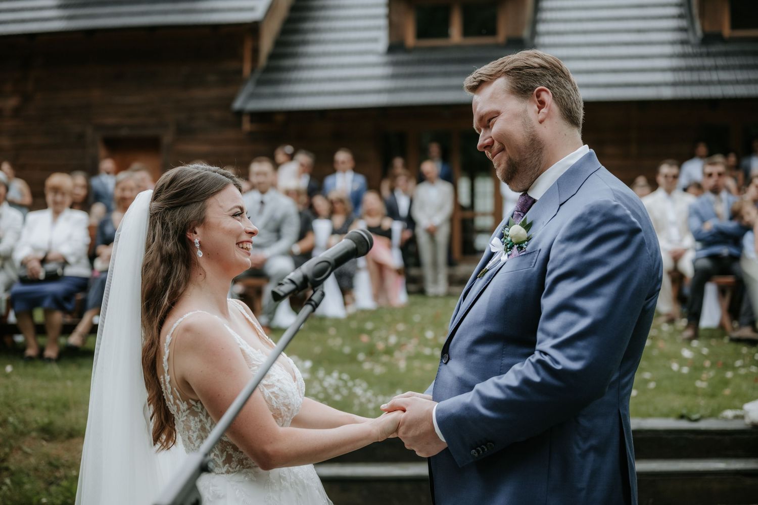 Bride and groom exchanging vows outdoors at rustic wedding ceremony with guests seated in background.