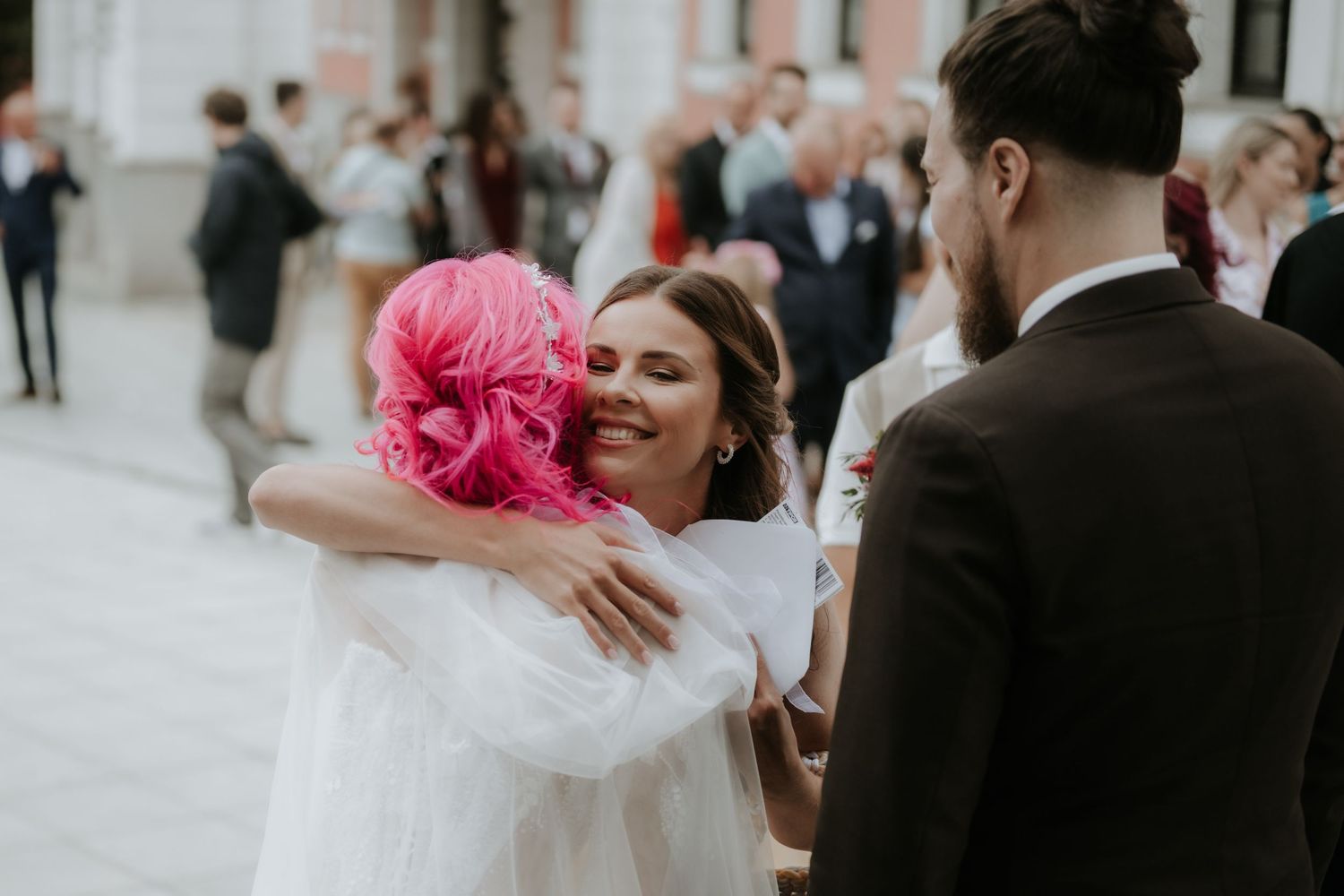 Bride in white dress hugging guest with pink hair at outdoor wedding reception.