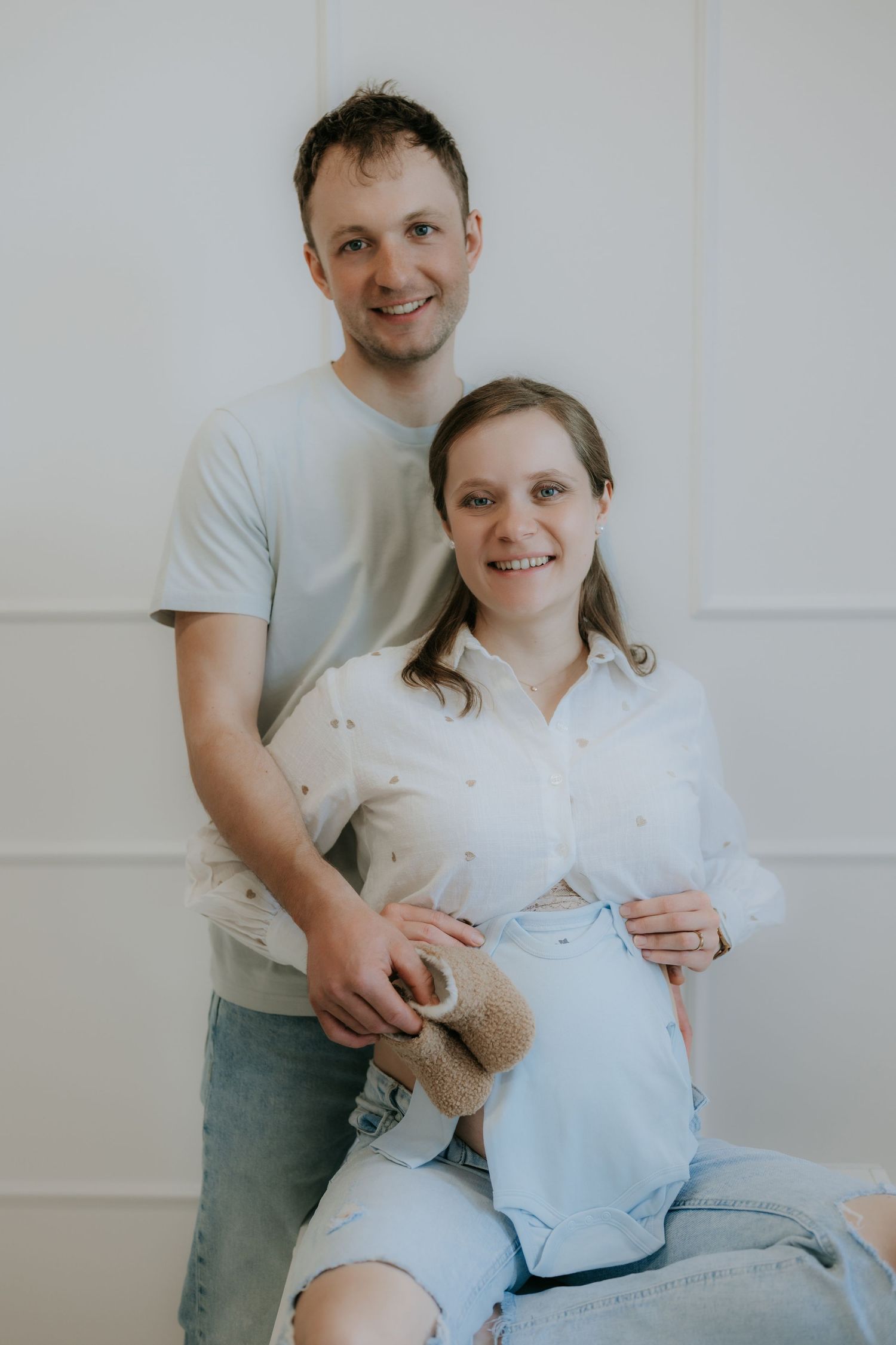 Smiling couple posing for maternity photo, man standing behind pregnant woman holding baby clothes.