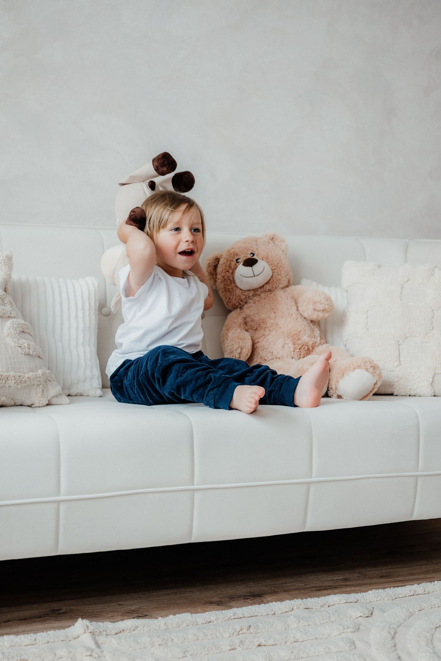 A cheerful toddler boy sits on a white sofa holding a small stuffed animal beside a large teddy bear.
