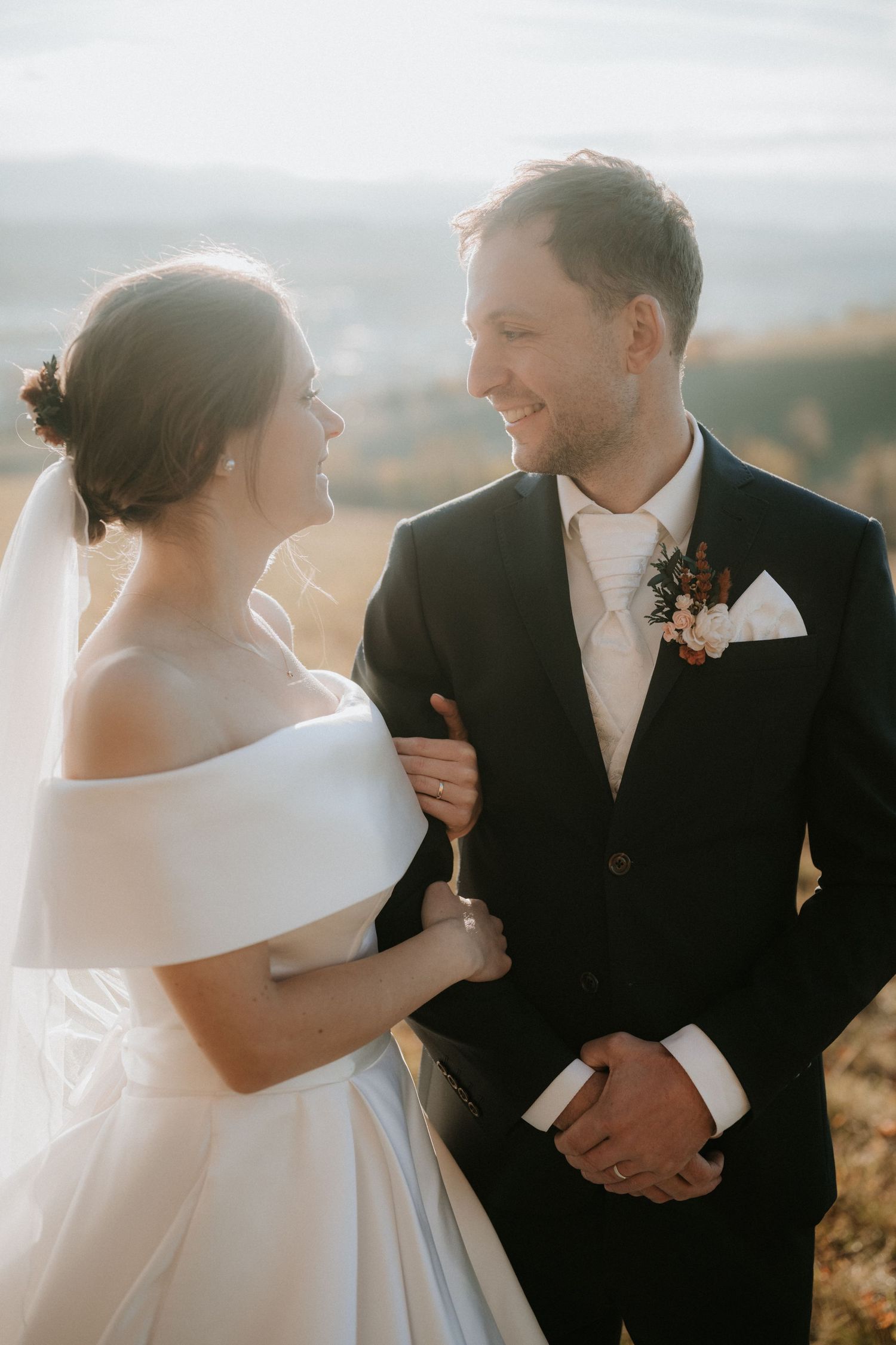 Bride in white off-shoulder gown and veil gazing at smiling groom in black suit on a sunny hillside.
