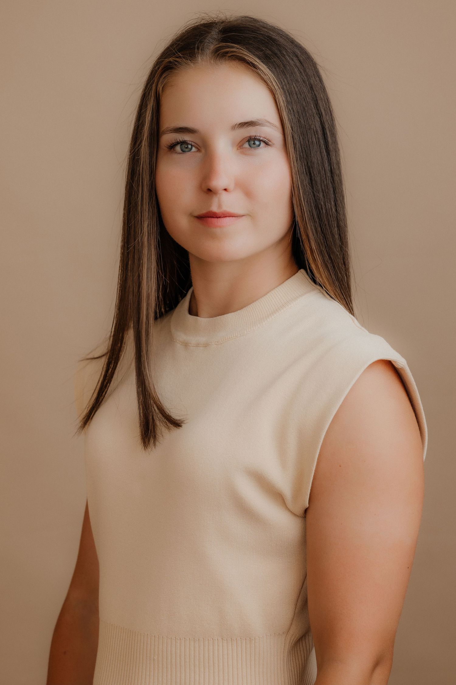 Young woman with straight brown hair wearing cream sleeveless ribbed top posing against beige background.