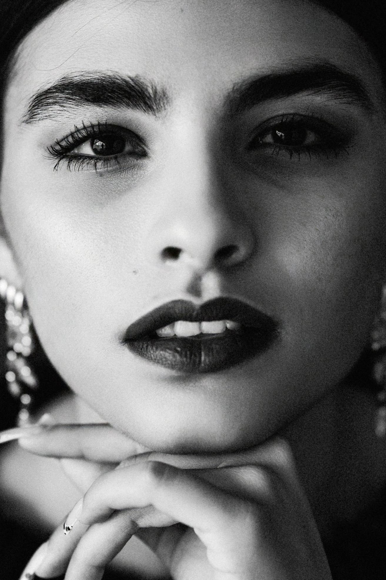 Black and white close-up of a young woman with bold brows, dark lips, and hoop earrings resting chin on hand.