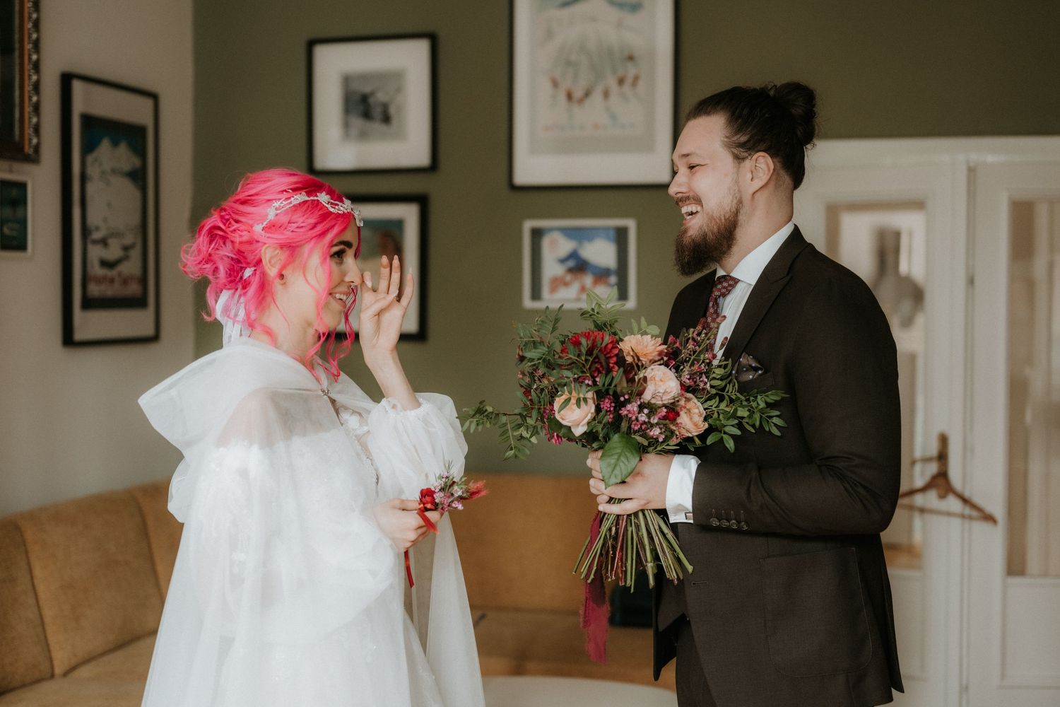 Bride with pink hair in white dress reacts emotionally as groom in dark suit presents a bridal bouquet indoors.
