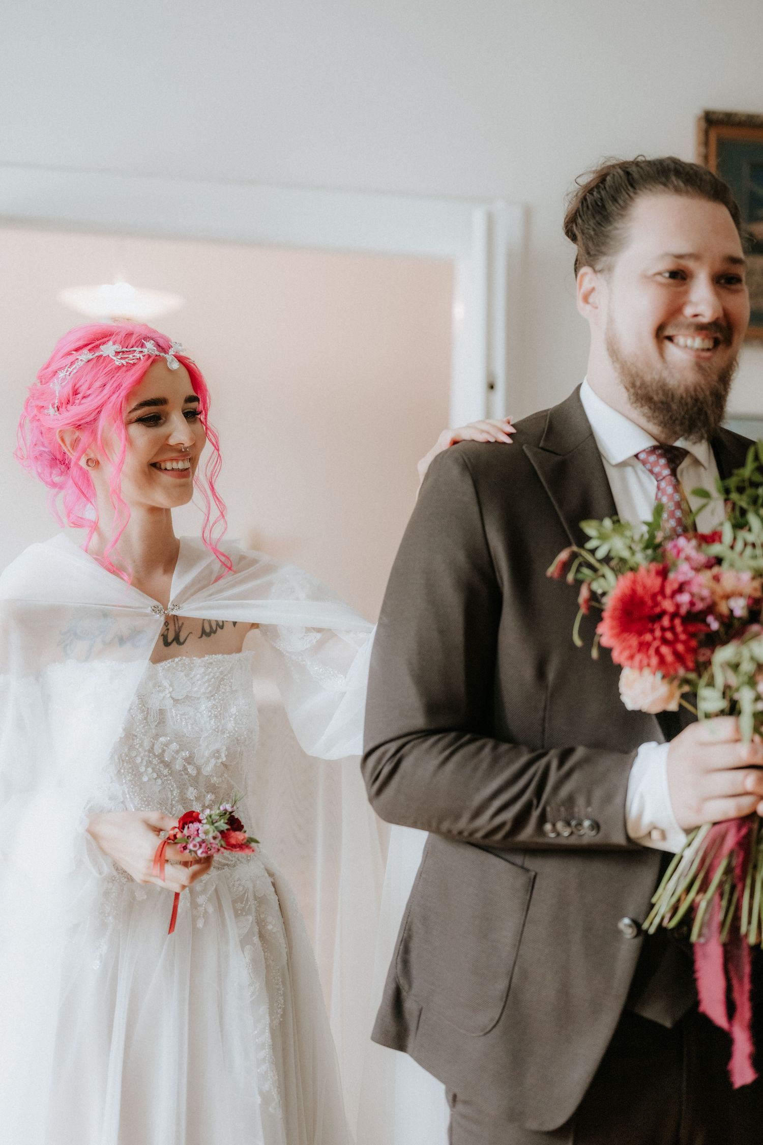 Bride with pink hair in white gown and groom in gray suit smiling and holding colorful bouquet at wedding ceremony.