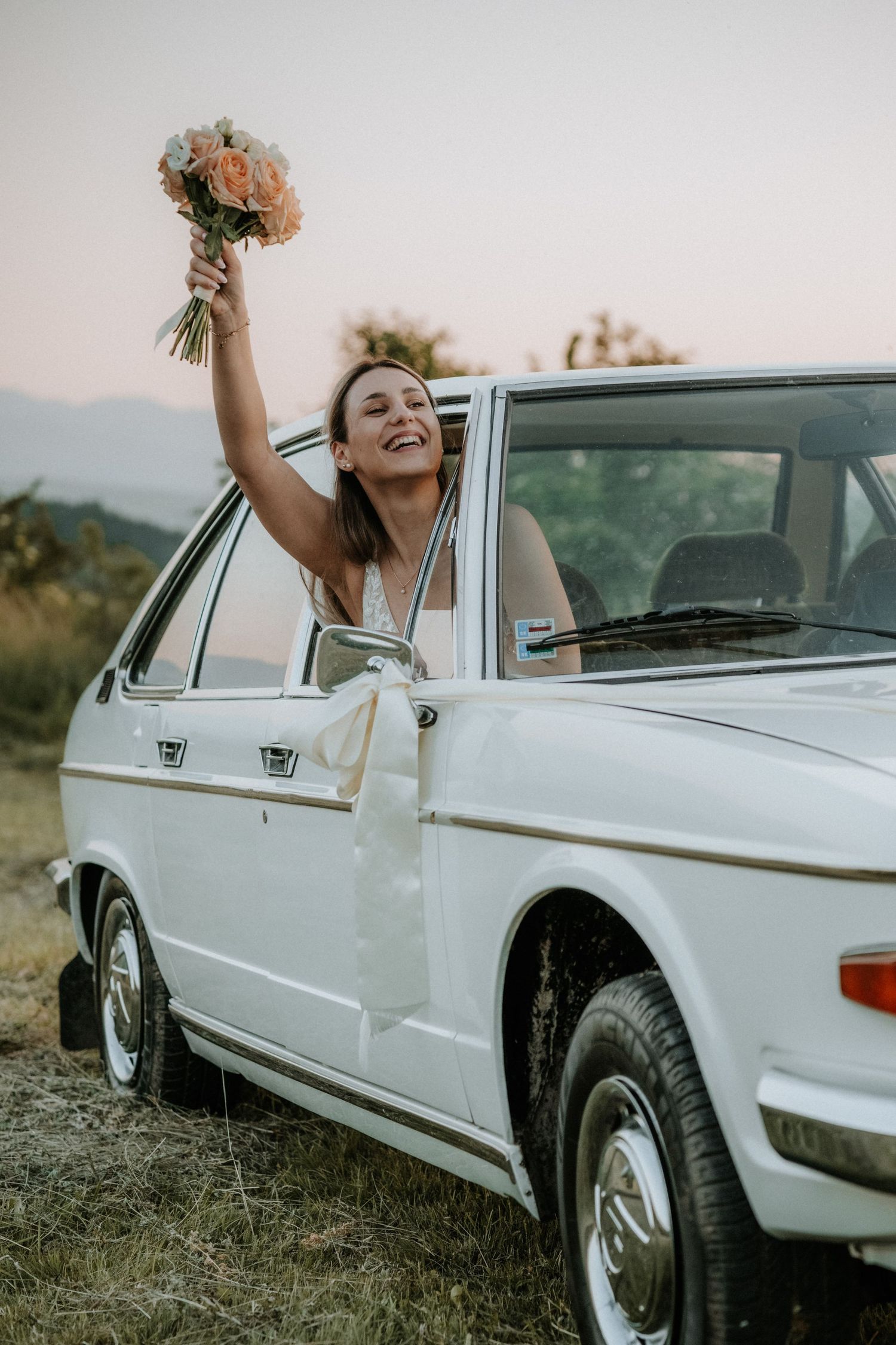 Joyful bride in white dress leaning out of vintage white car, raising bouquet of flowers against sunset sky.